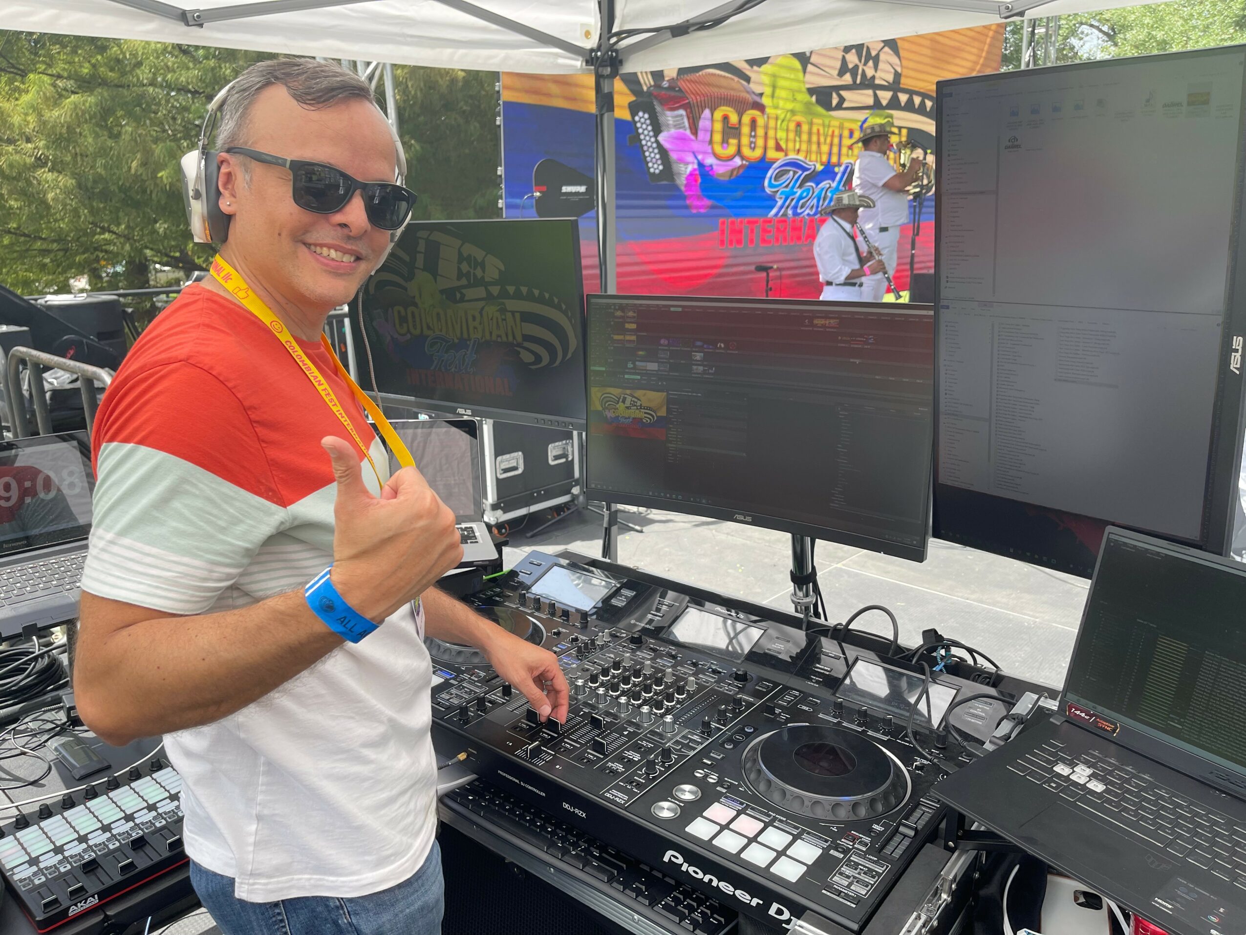 Man wearing sunglasses and a red and white shirt, smiling at DJ equipment outdoors with screens and trees in background.