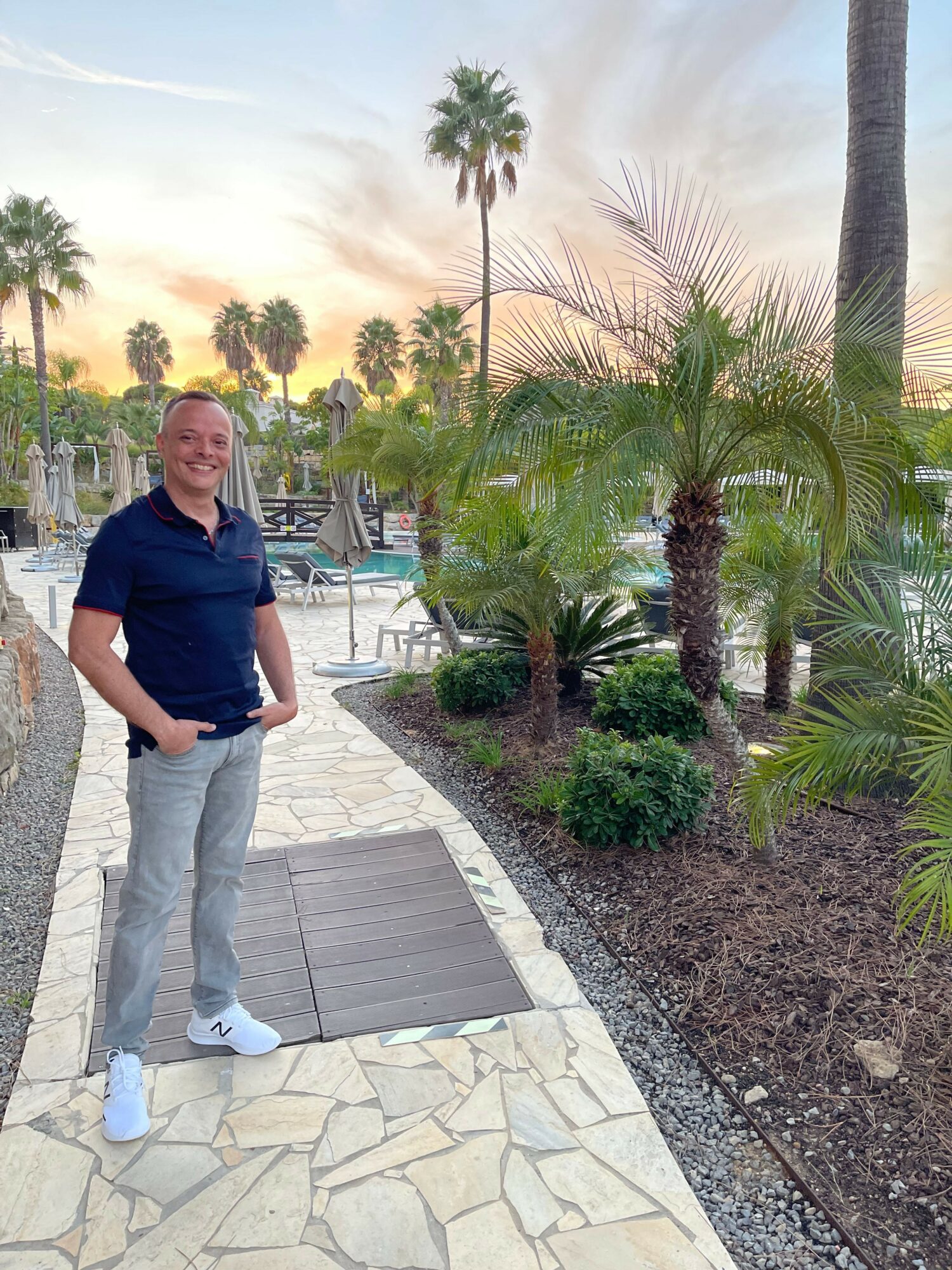 Man standing on a paved walkway with palm trees and a sunset sky in the background.