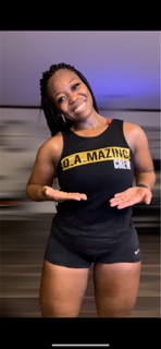 Smiling woman with braided hair in black athletic outfit, standing indoors, hands gesturing, background blurred.
