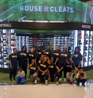 Group of nine people, some kneeling, in front of a shoe display at a store, with a sign reading 'HOUSE OF CLEATS'.
