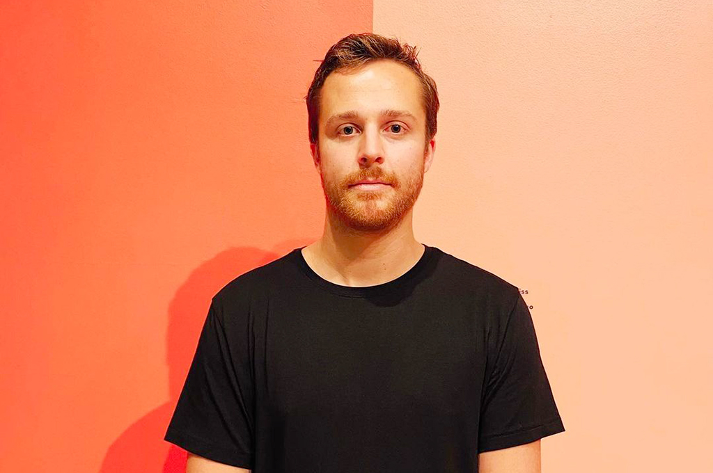 Young man with short hair and beard wearing a black t-shirt, standing against a pink background.