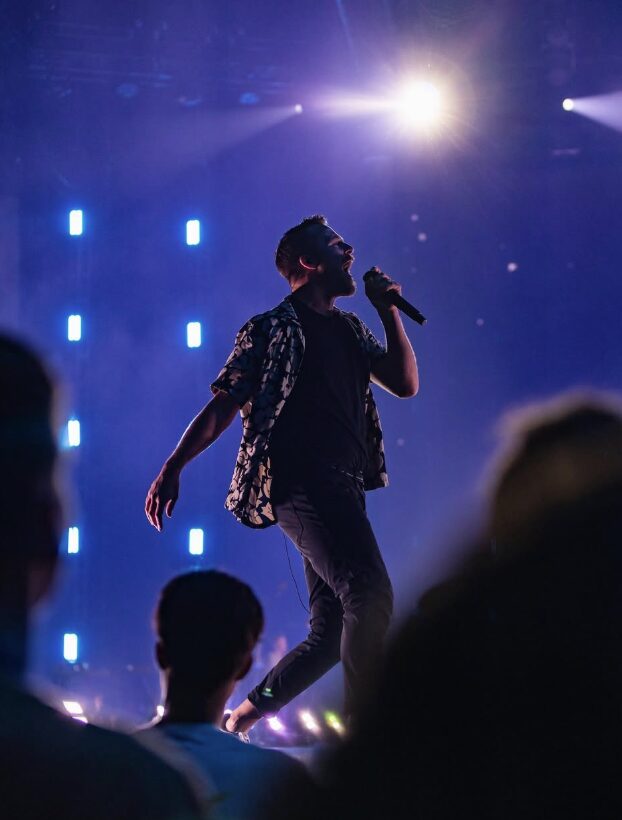Performer singing on stage with bright lights and audience silhouettes in foreground.