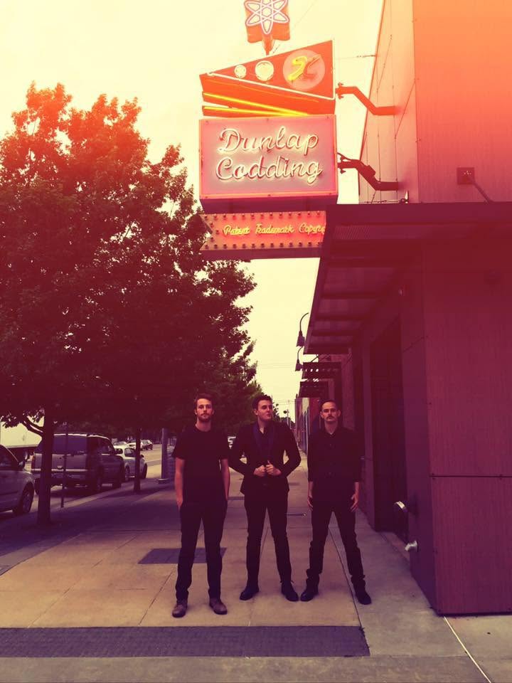 Three people stand on a sidewalk in front of a building with a sign and a tree nearby.