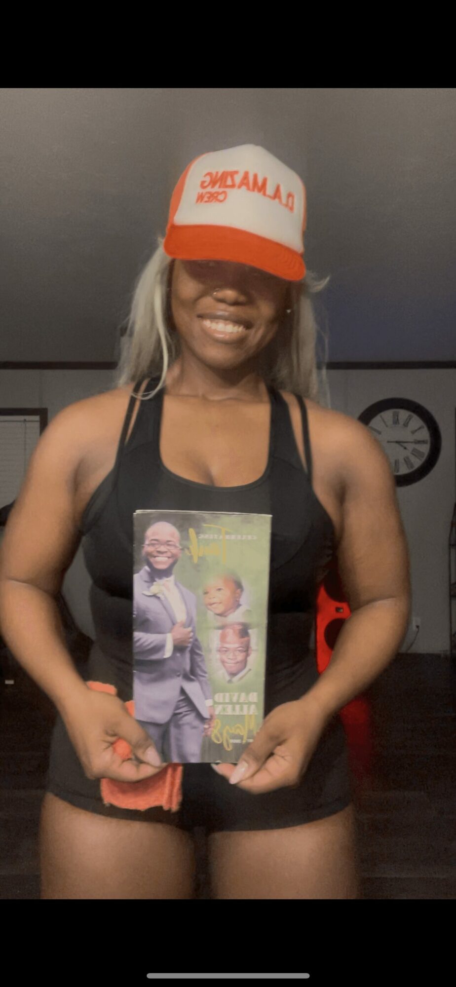 Woman wearing a white and orange cap, black tank top, holding a photo, smiling, in an indoor setting.