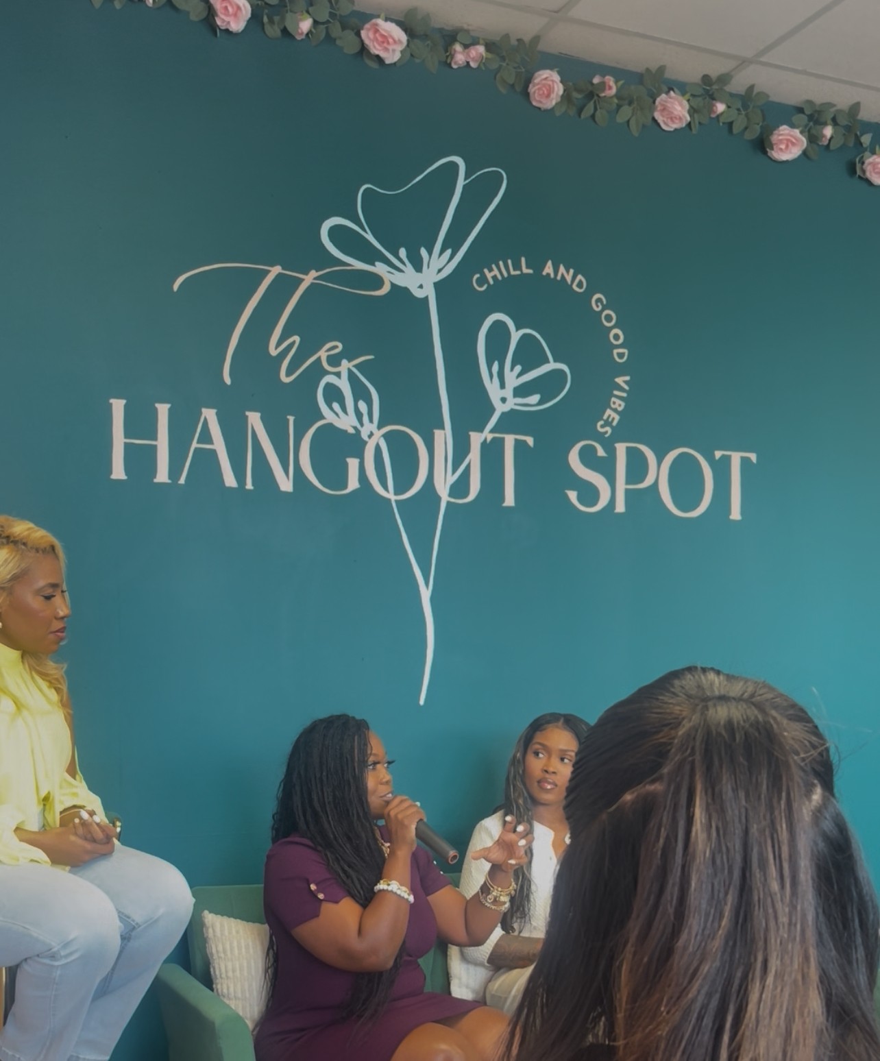 Group of women sitting and talking in front of a teal wall with a logo and text, floral decorations above.