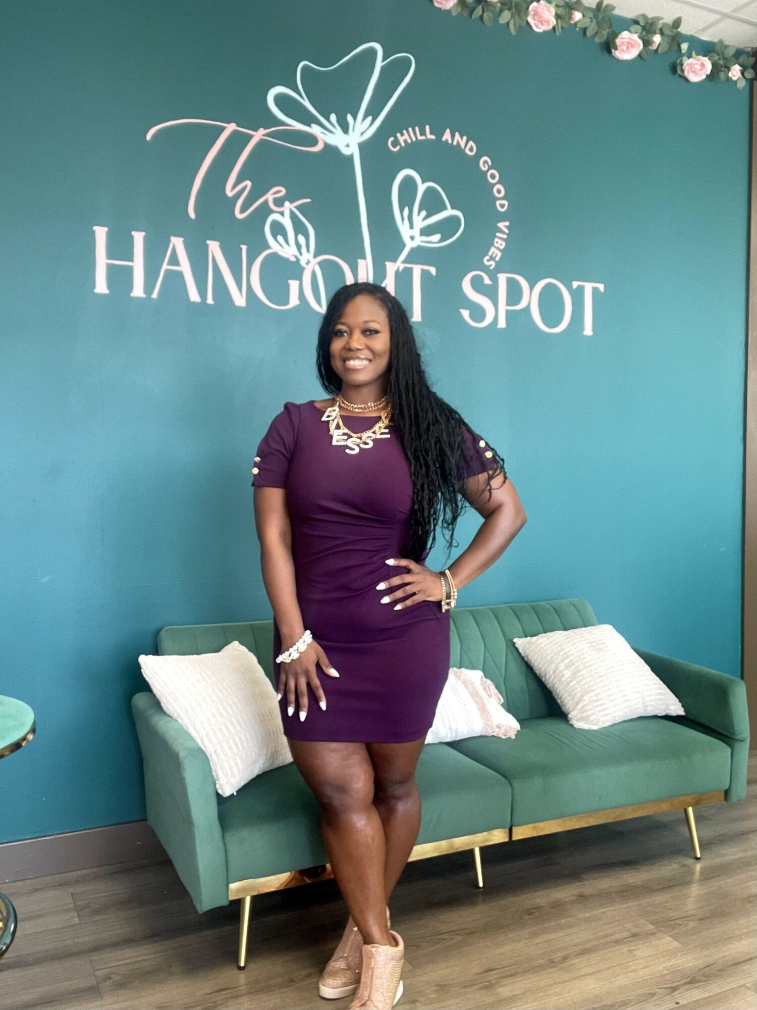 Woman in purple dress standing in front of teal wall with white text and floral decorations, smiling, with a green sofa and pillows behind her.