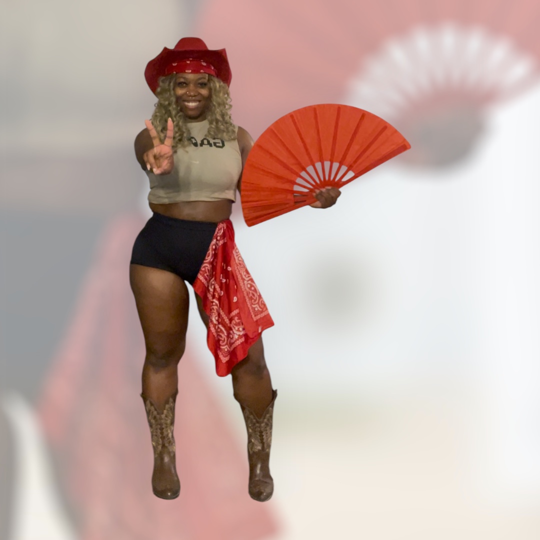 Person with a red hat, holding a red fan, smiling, making a peace sign, wearing shorts and a bandana, standing against a blurred background.