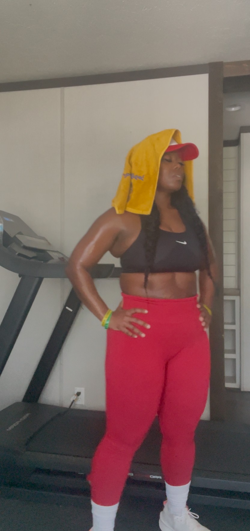 Woman standing on treadmill with towel on her head, wearing a black top and red pants, in a gym.