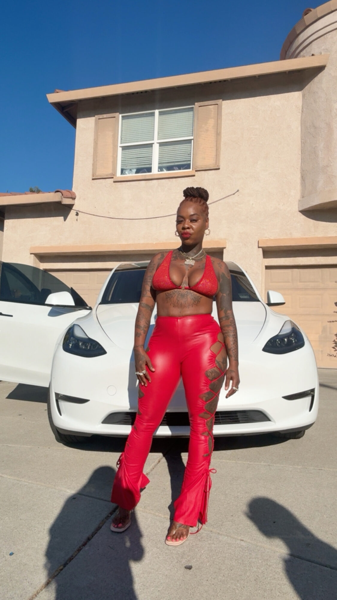Woman in red outfit standing in front of a white car outside a beige house with a blue sky.