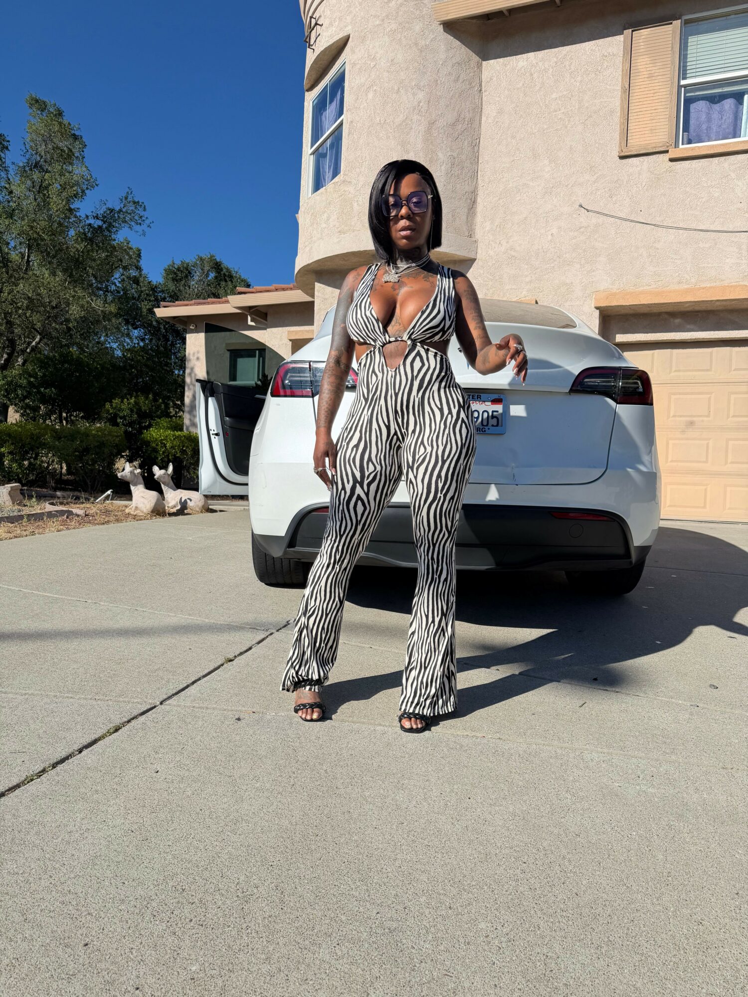 Woman in striped jumpsuit standing next to a silver car in a parking lot, with buildings and trees in background.