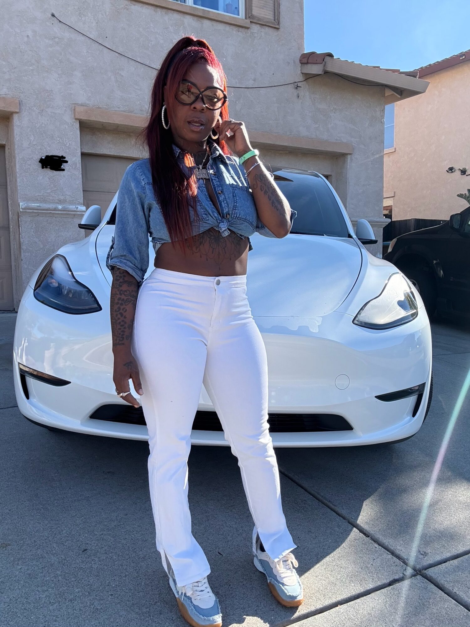 Woman with long hair, glasses, and tattoos standing in front of a white sports car, wearing a denim jacket and white pants.