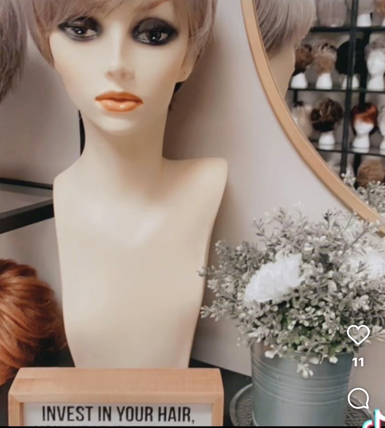 Mannequin head with styled hair, next to a flower arrangement in a pot, in a store setting.