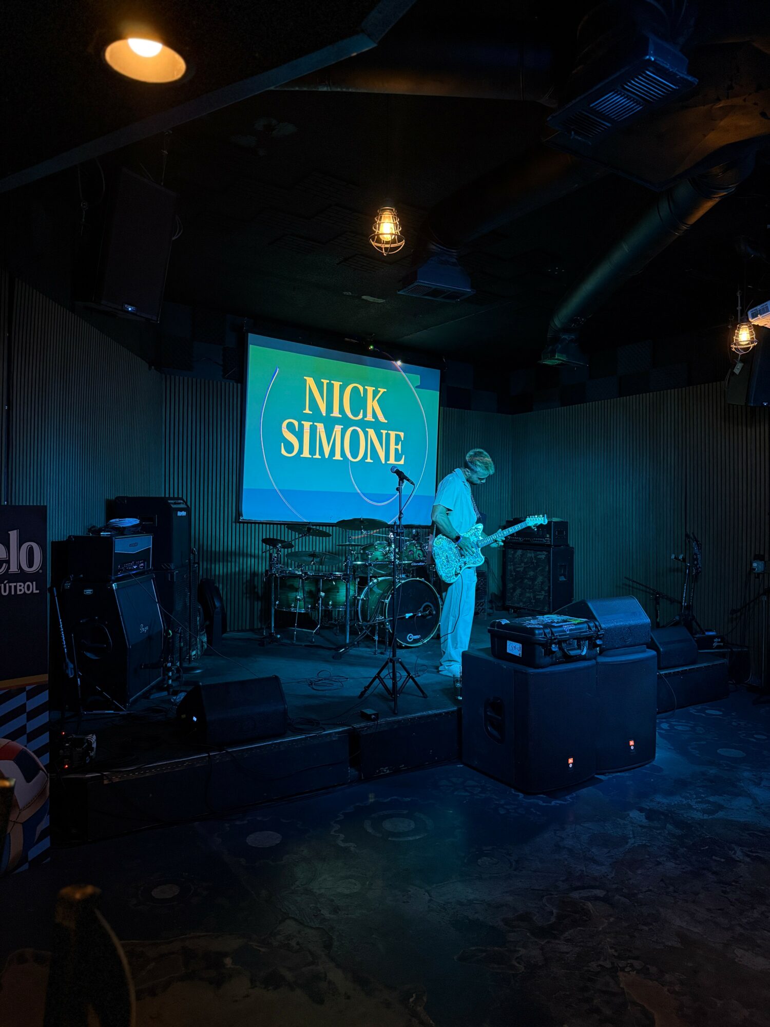 Musician playing guitar on stage with a screen displaying 'NICK SIMONE' behind him.