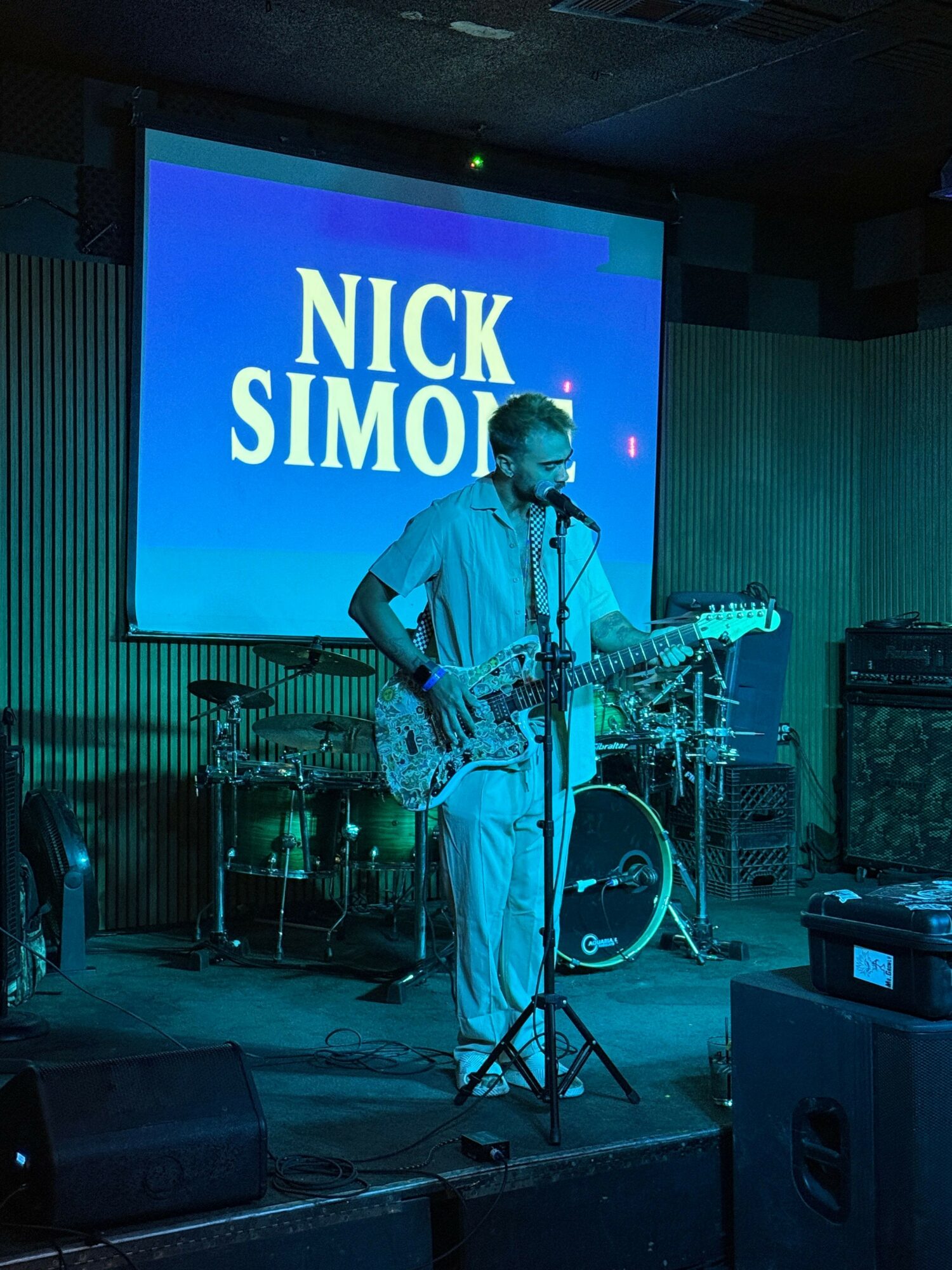 Person on stage with microphone, large screen behind displaying 'NICK SIMO', stage equipment visible.