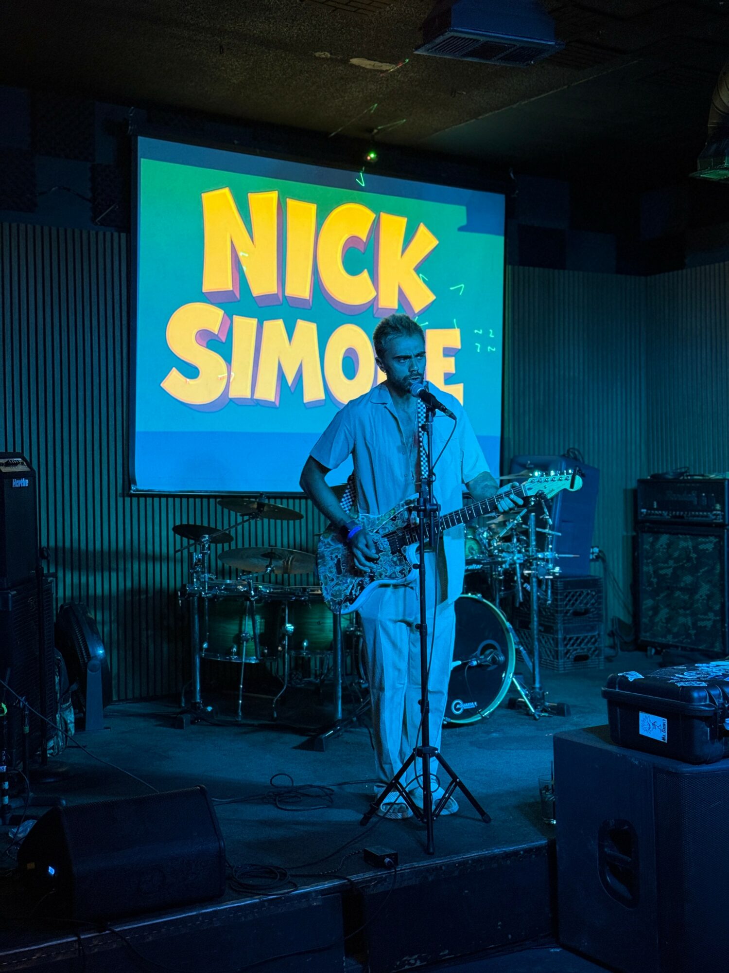 Musician playing guitar on stage with a large screen displaying 'NICK SIMONE' behind him.