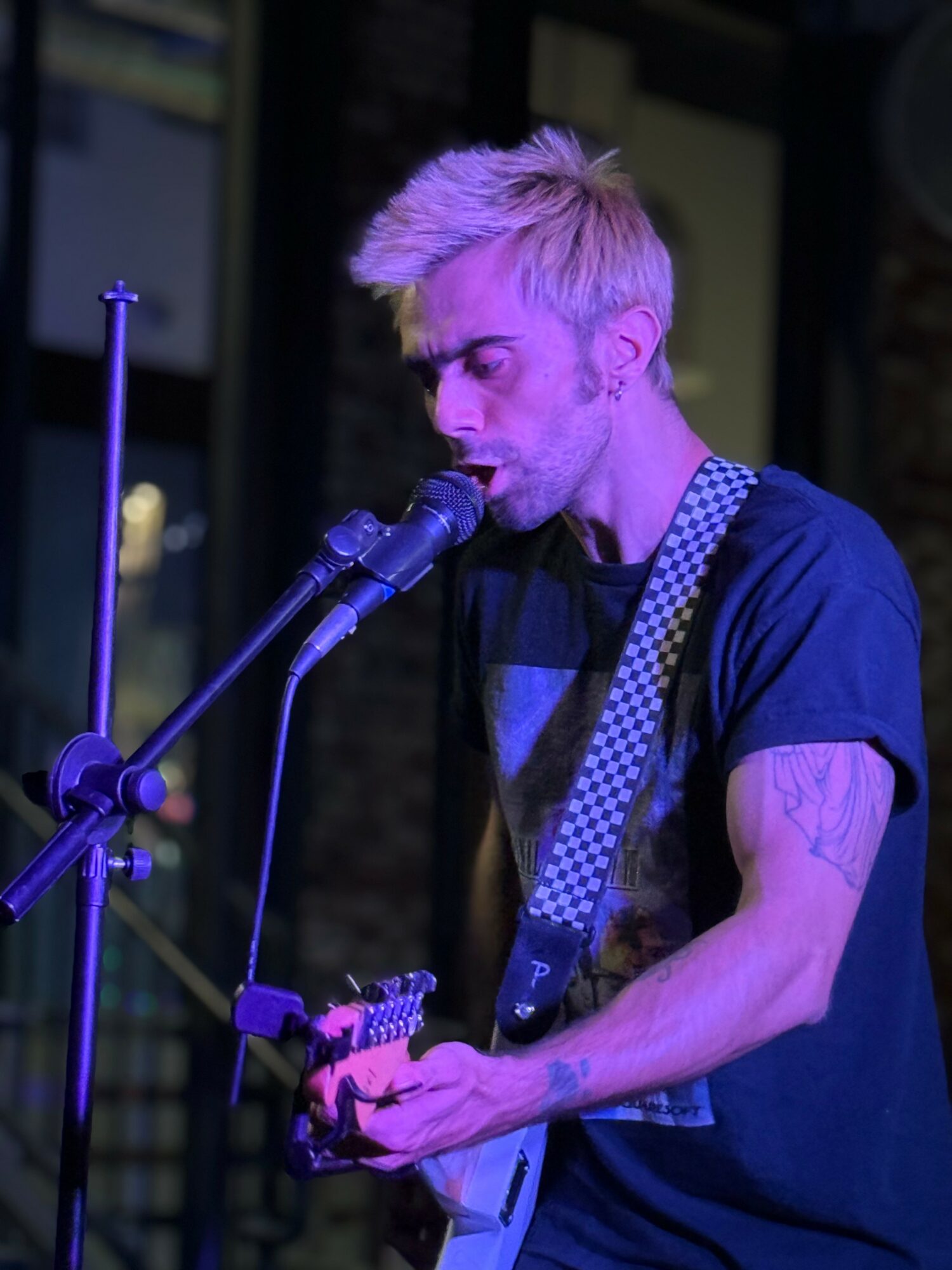 Young man with blonde hair singing into a microphone while playing guitar, wearing a dark t-shirt and patterned strap.