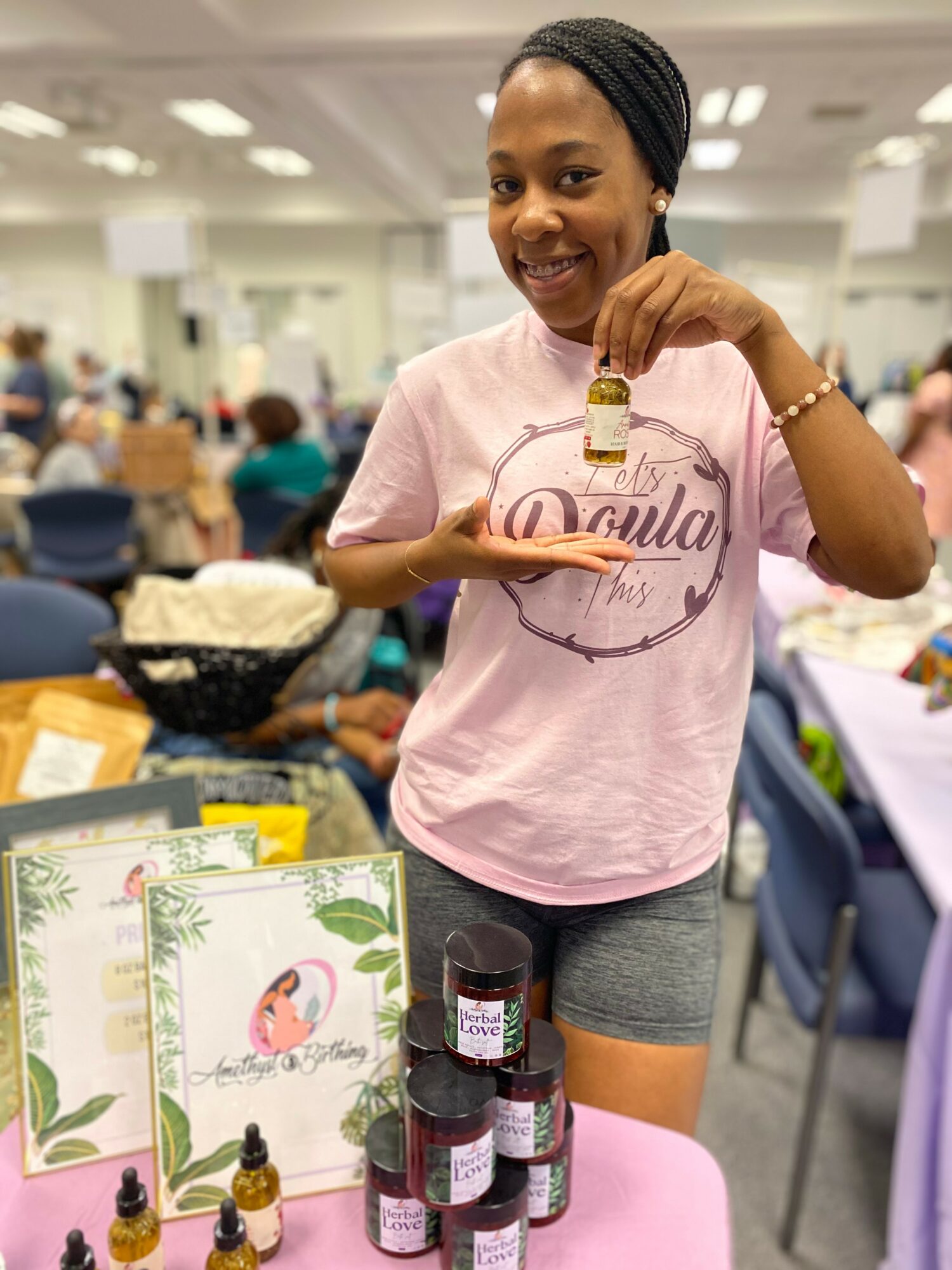 Smiling girl holding a small bottle in a large indoor event space with tables and people, standing next to a pink table with products.