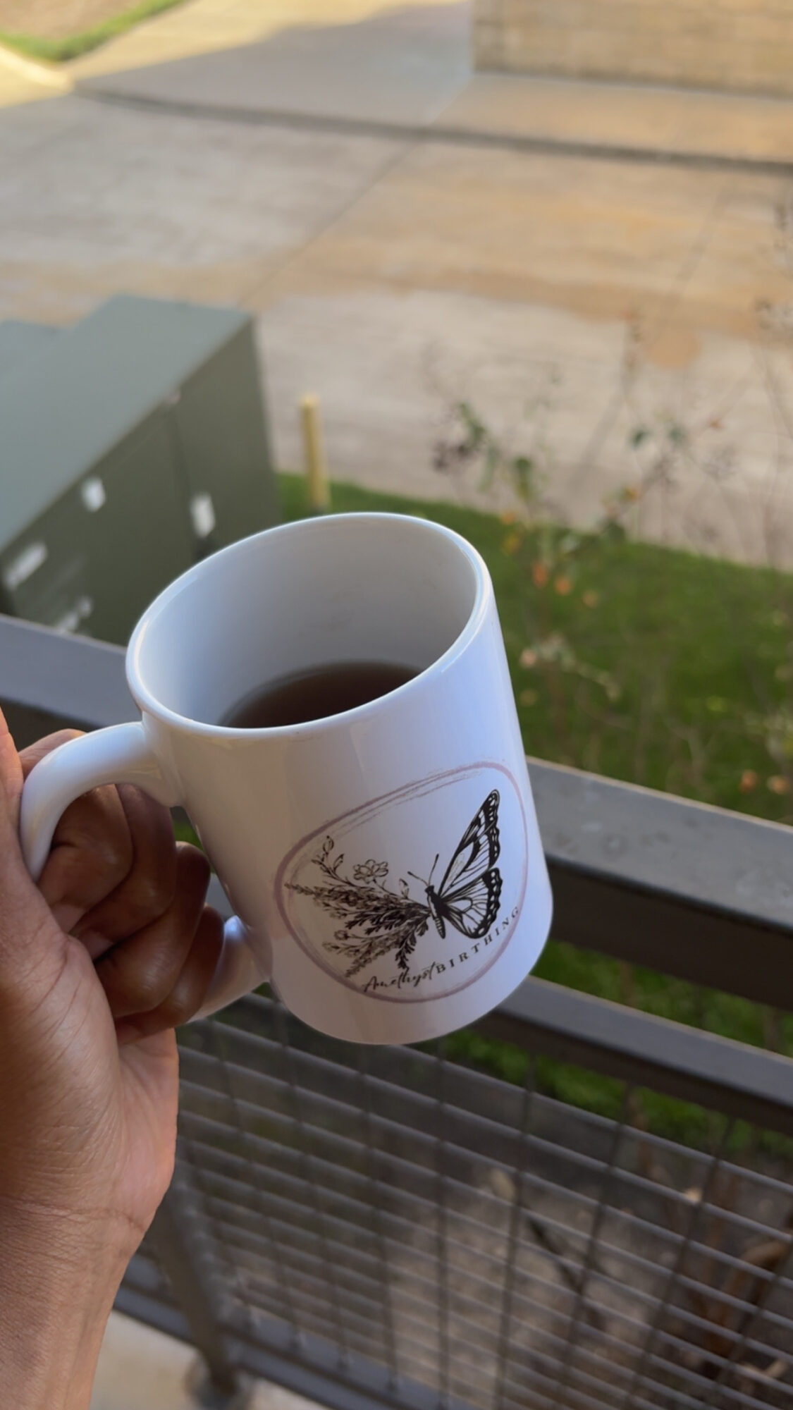 Person holding a white mug with a butterfly and plant design, filled with dark liquid, outdoors on a balcony railing.