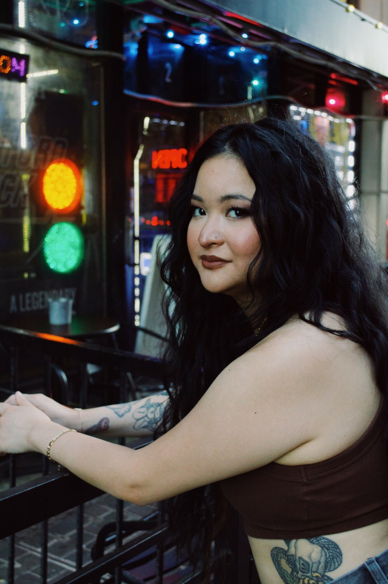 Young woman with long dark hair and tattoos, sitting at a bar or counter, with colorful lights and signs in the background.