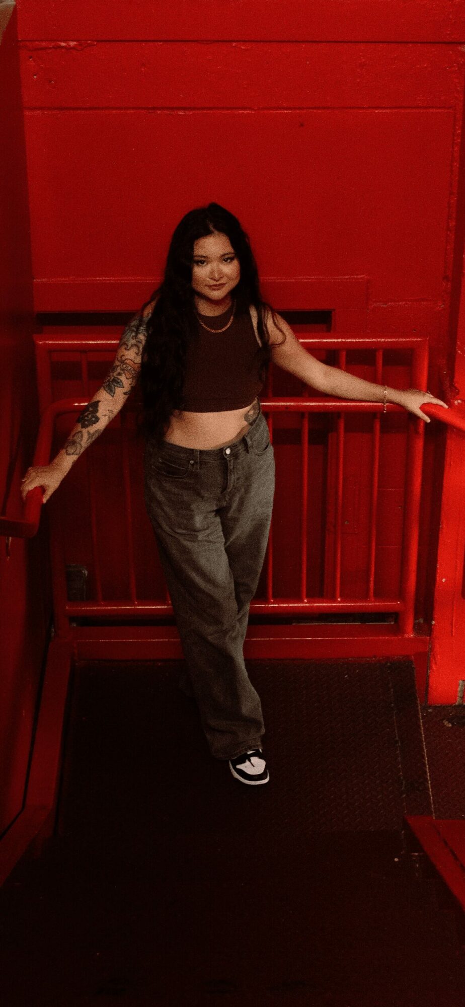 Woman with long dark hair and tattoos on her arms, wearing a black crop top and gray pants, standing in front of a red wall.