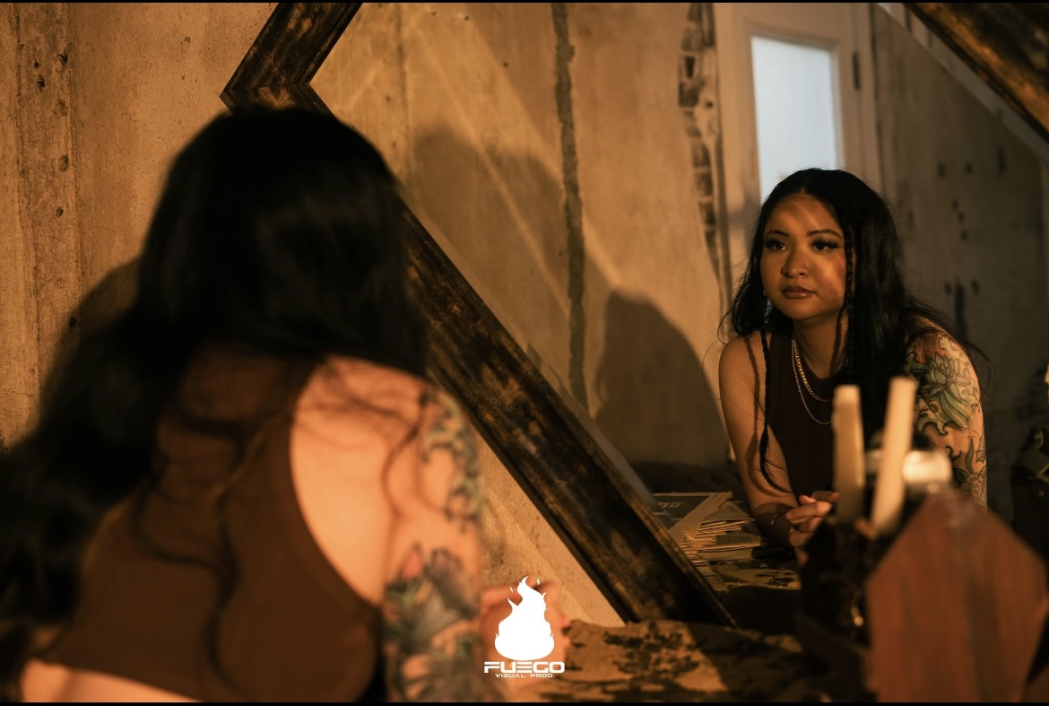 Two women with tattoos sit at a table in a dimly lit room, facing each other, with candles and rustic decor.