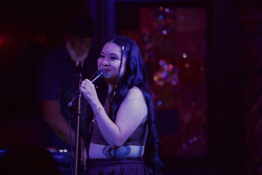 A woman with long dark hair holding a microphone, smiling, in a dimly lit setting with a person in the background.