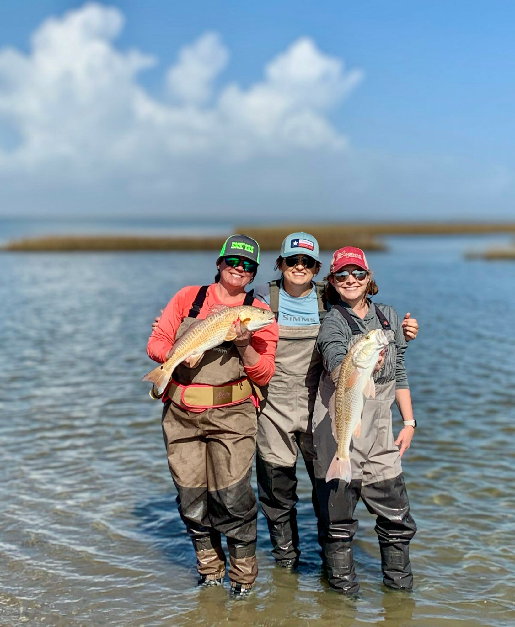 Three people standing in shallow water holding fish, wearing waders and hats, with a cloudy sky and water in background.