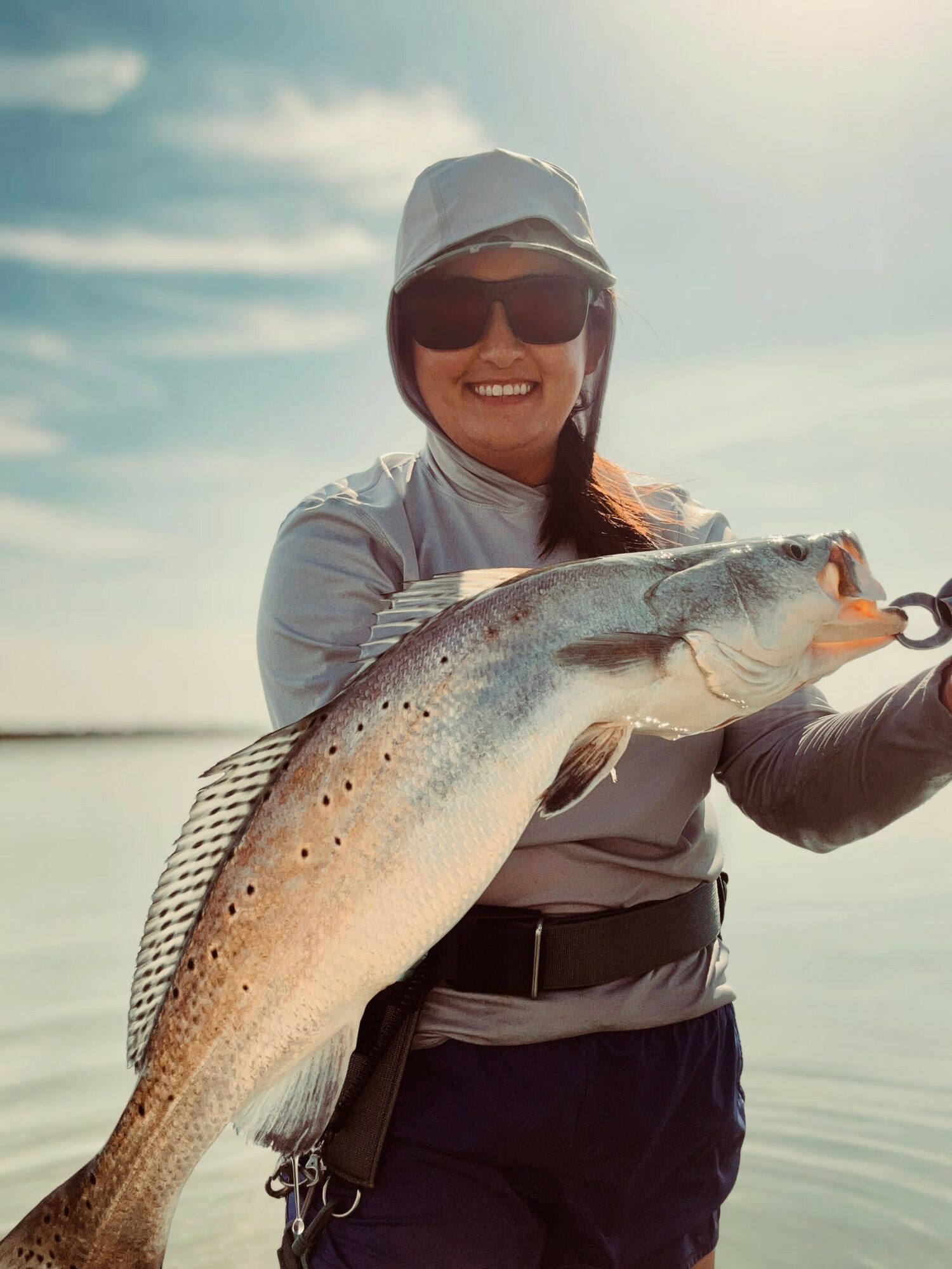 Smiling woman holding a large fish outdoors with water and sky in background.