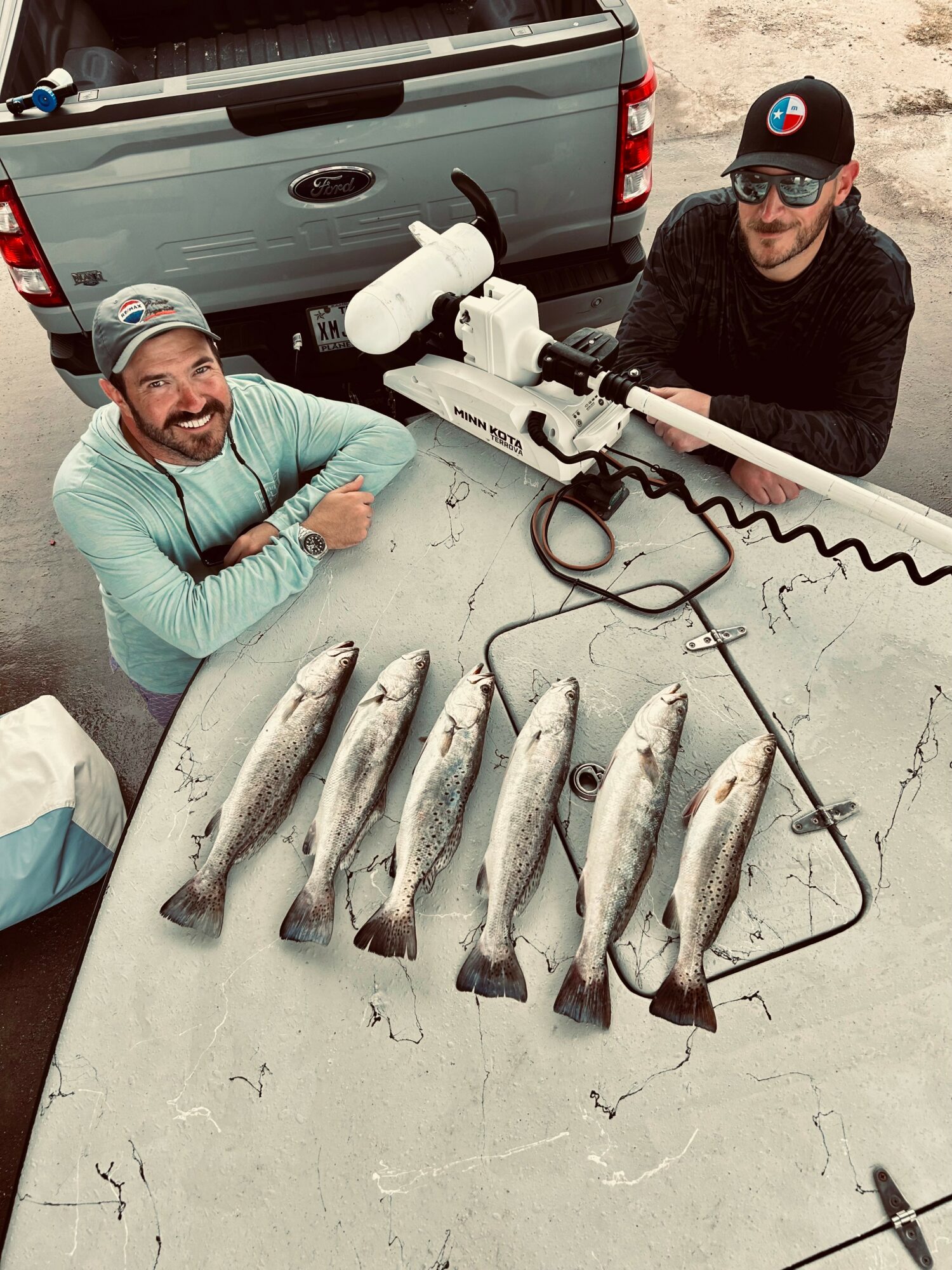 Two men smiling at a table with seven fish, a fishing gun, and fishing tools outdoors near a truck.