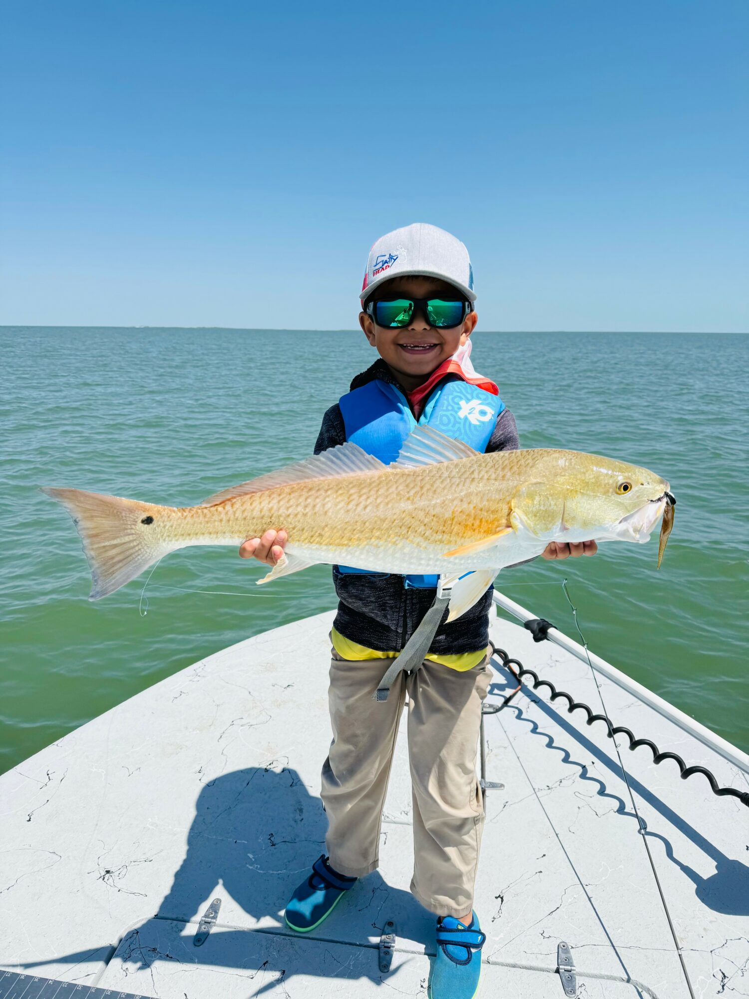 Child in sunglasses and hat holding a large fish on a boat, with water and sky in background.