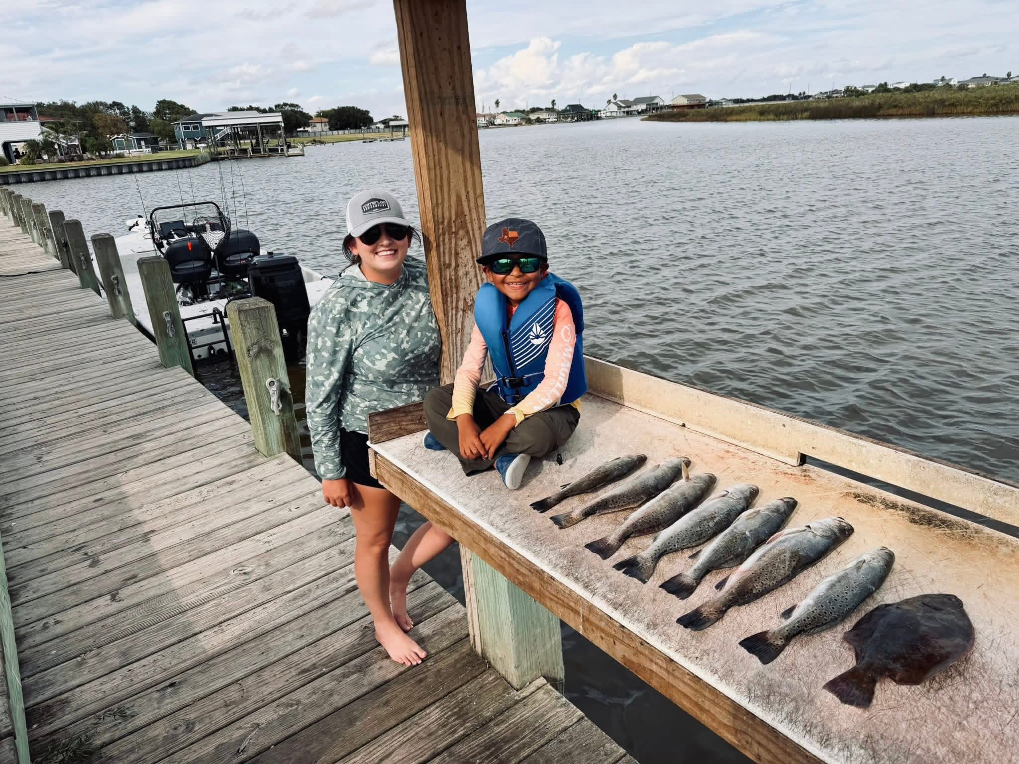 Two children stand on a wooden dock near water, with a display of fish laid out on a wooden table.