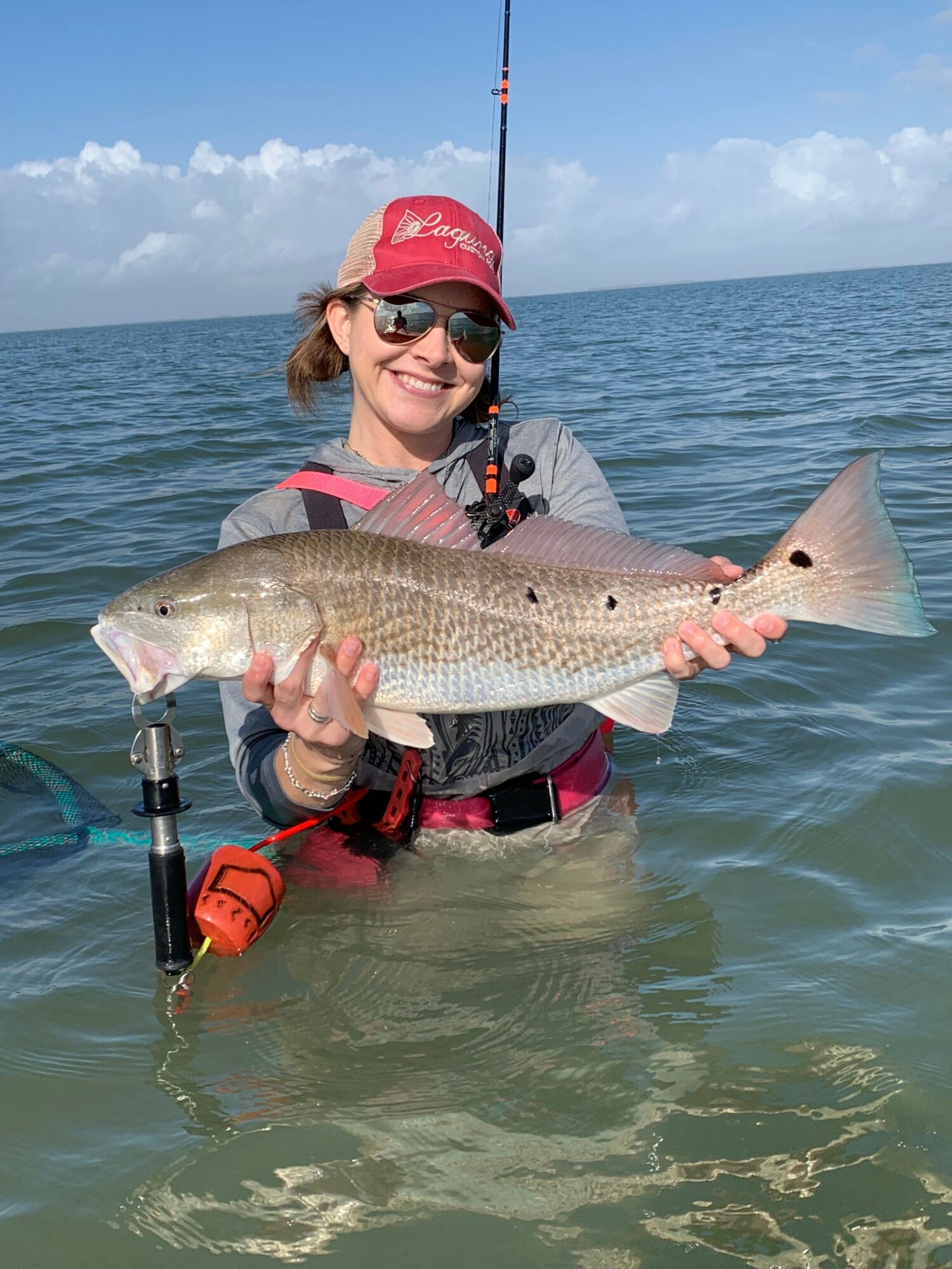 Person holding a large fish while standing in water, wearing sunglasses and a red cap, with a fishing rod nearby.