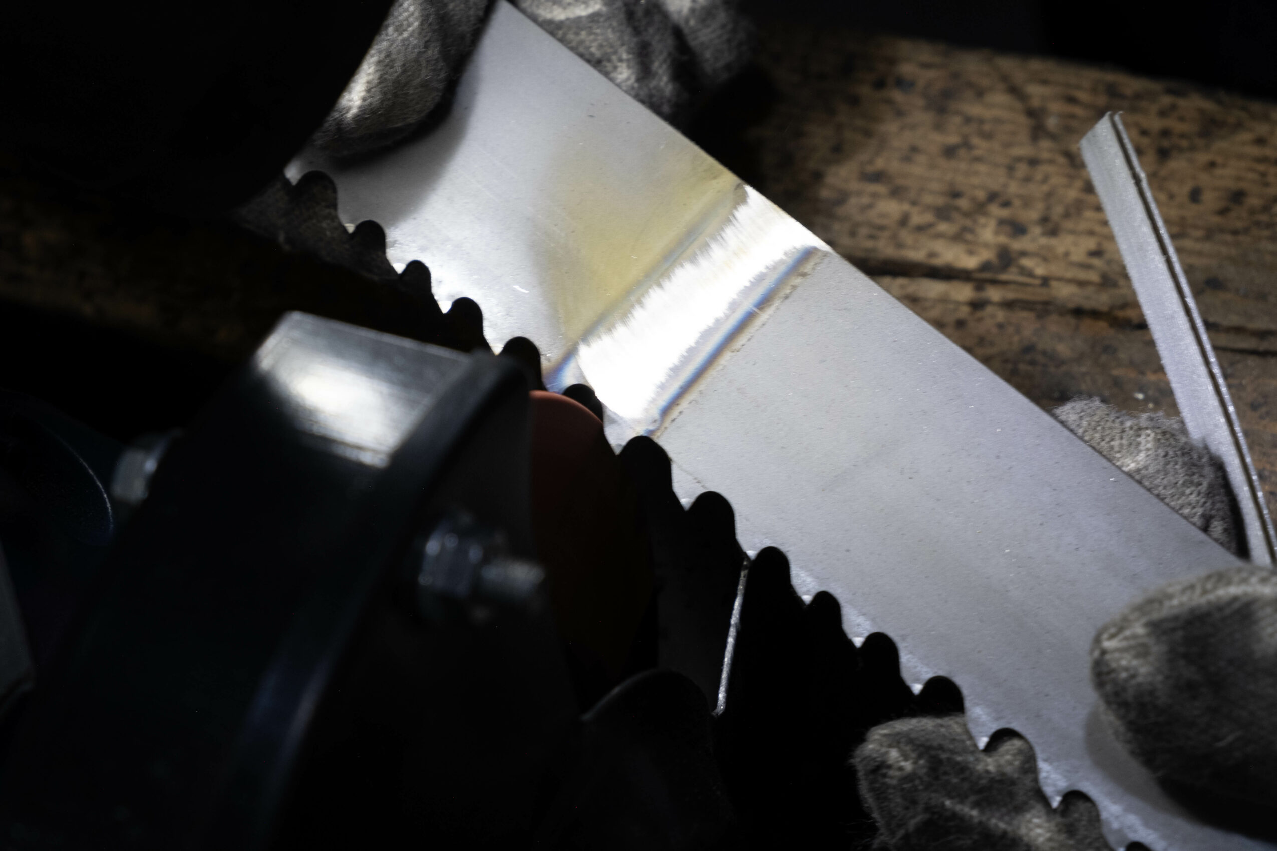 Saw blade with jagged edge on a wooden surface, partially covered by a gloved hand, with a metal ruler nearby.