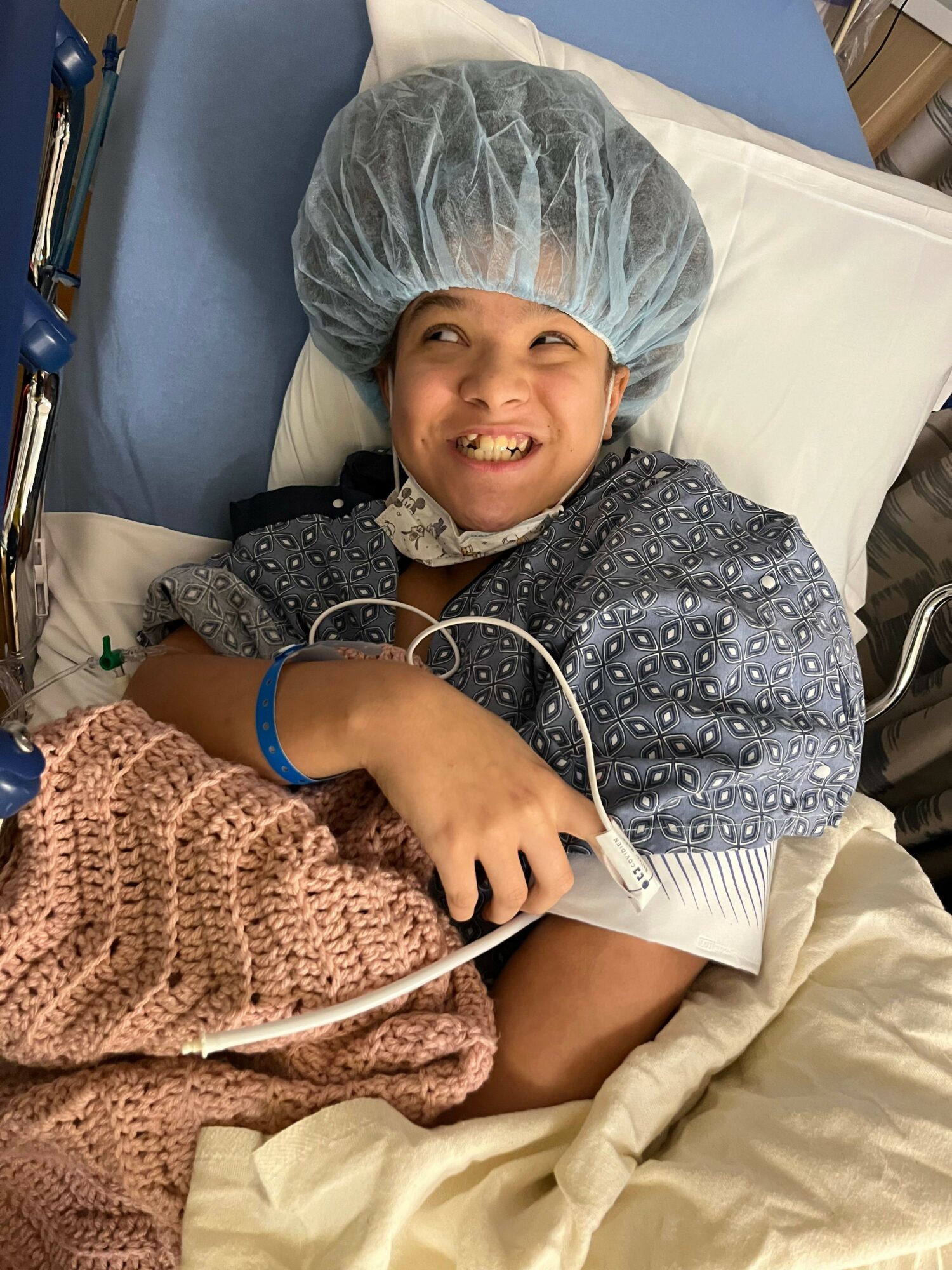 Child lying in hospital bed with a medical cap, smiling, connected to a monitor, covered with a blanket.