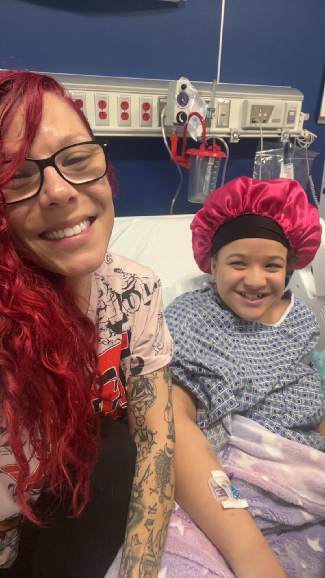 Woman with red hair and glasses smiling next to a young girl in hospital bed wearing a pink cap, smiling.