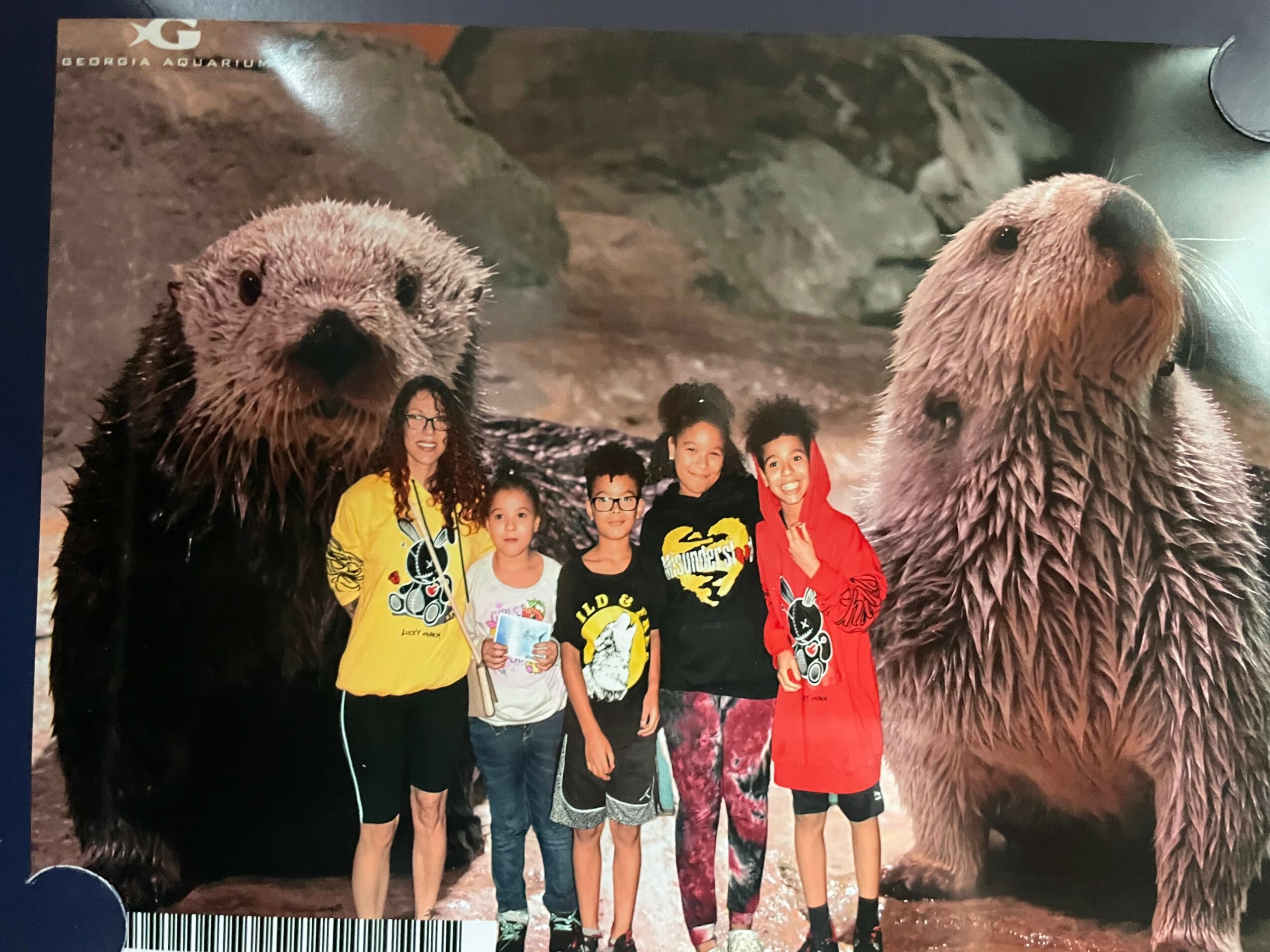 Five children stand in front of a large backdrop featuring two otters, one on each side, with rocky scenery in the background.