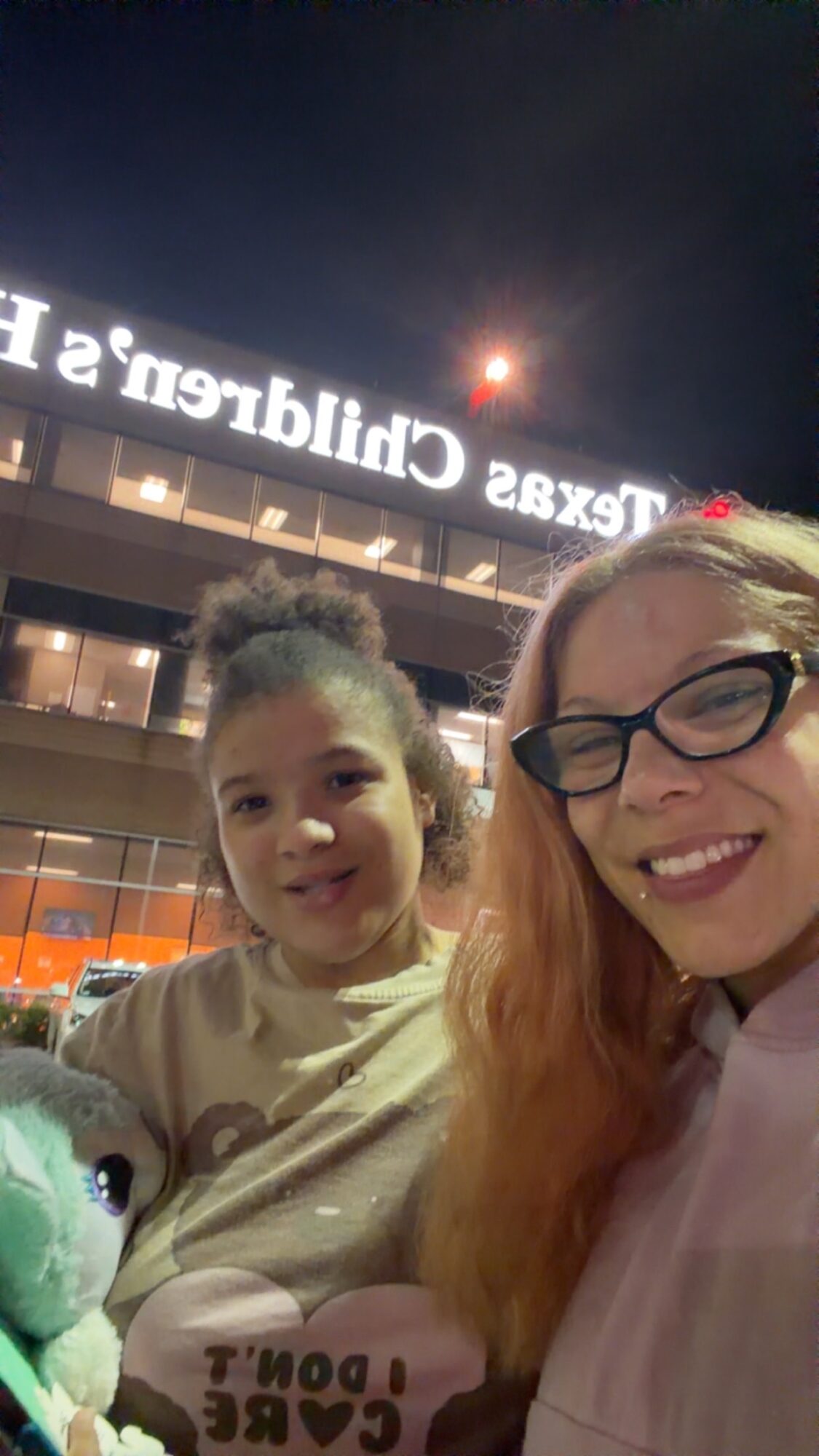Two people smiling outdoors at night, with a building and illuminated sign in the background.