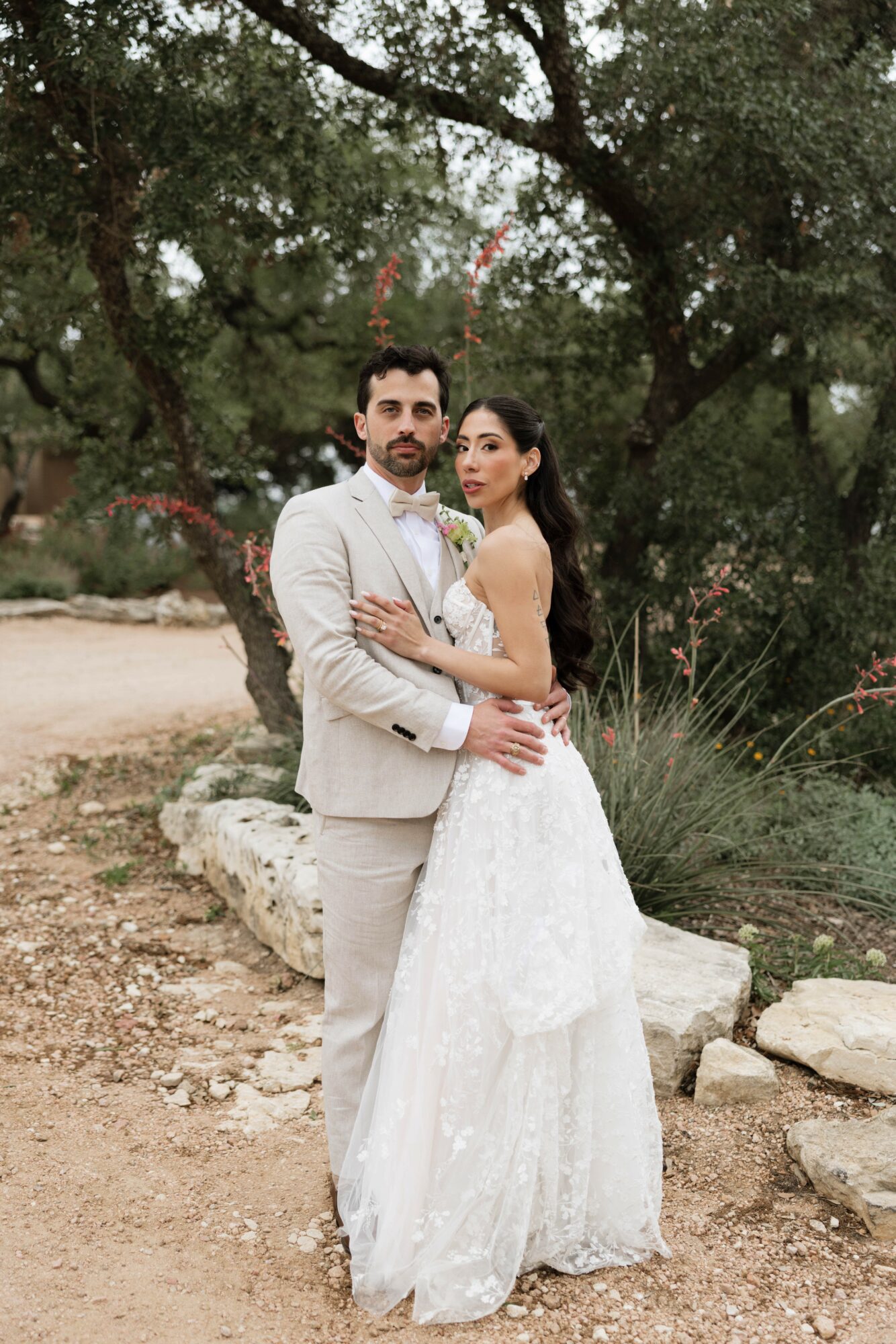 A couple in wedding attire standing outdoors among trees and rocks, embracing each other.