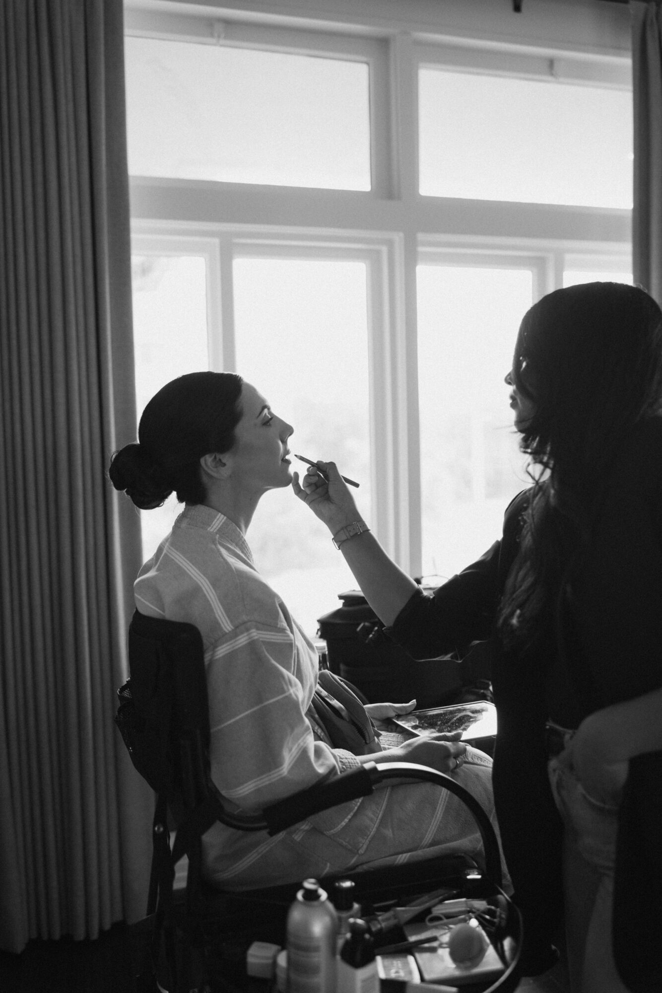 Person applying makeup to another person sitting in a chair near a large window, silhouette style.