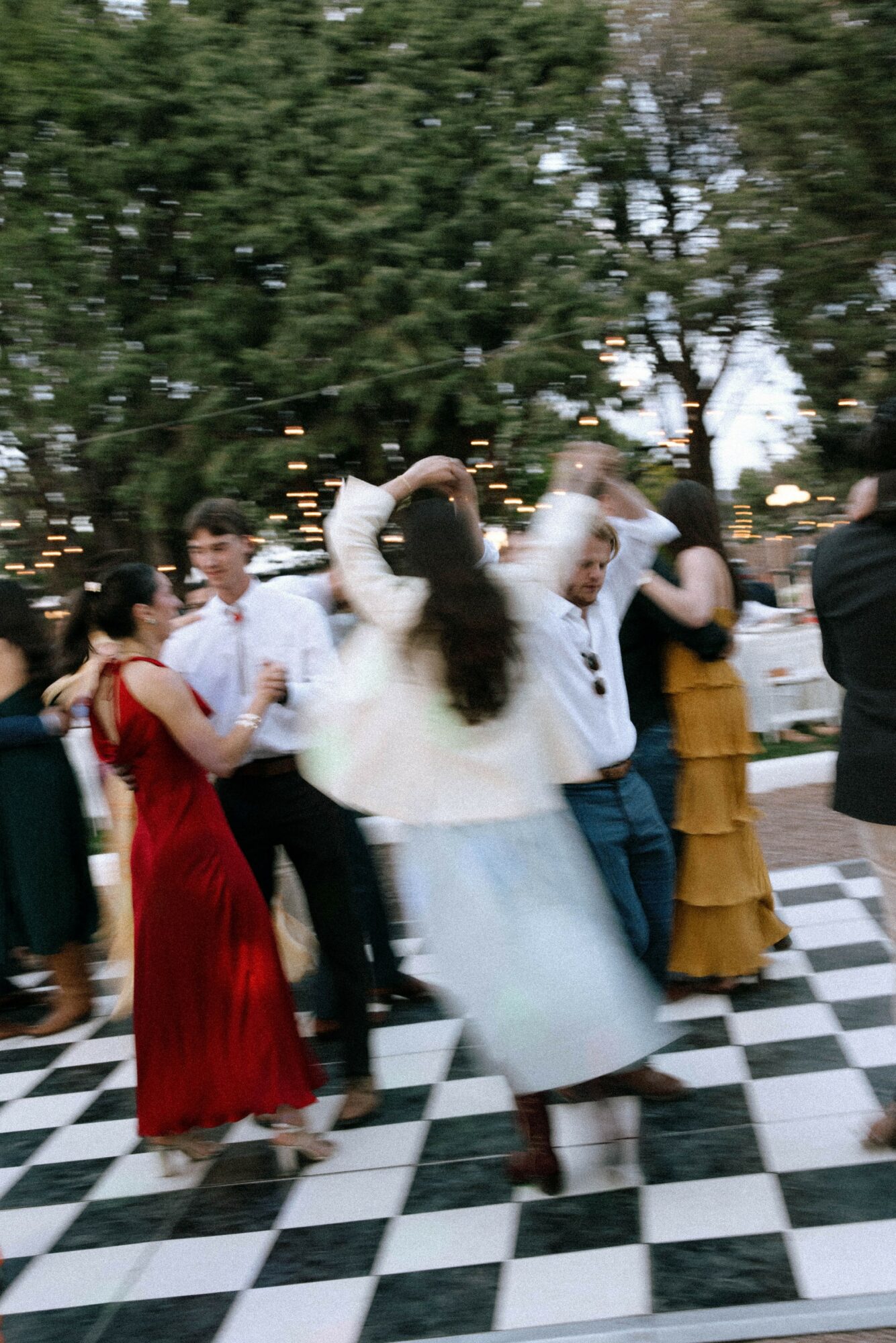 People dancing outdoors on a checkered floor, surrounded by trees, with some wearing formal attire, during evening time.