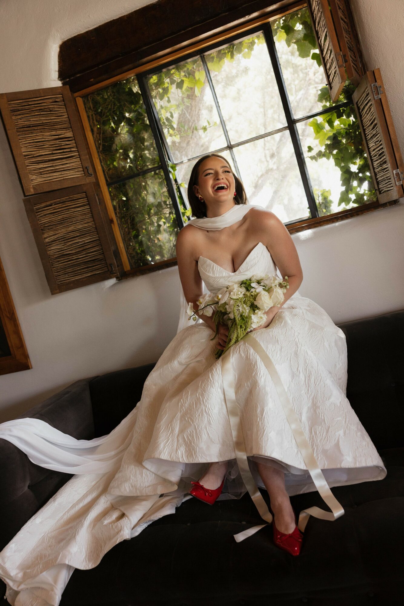 Woman in wedding dress sitting on a sofa, holding a bouquet, laughing, with open window behind her showing greenery.