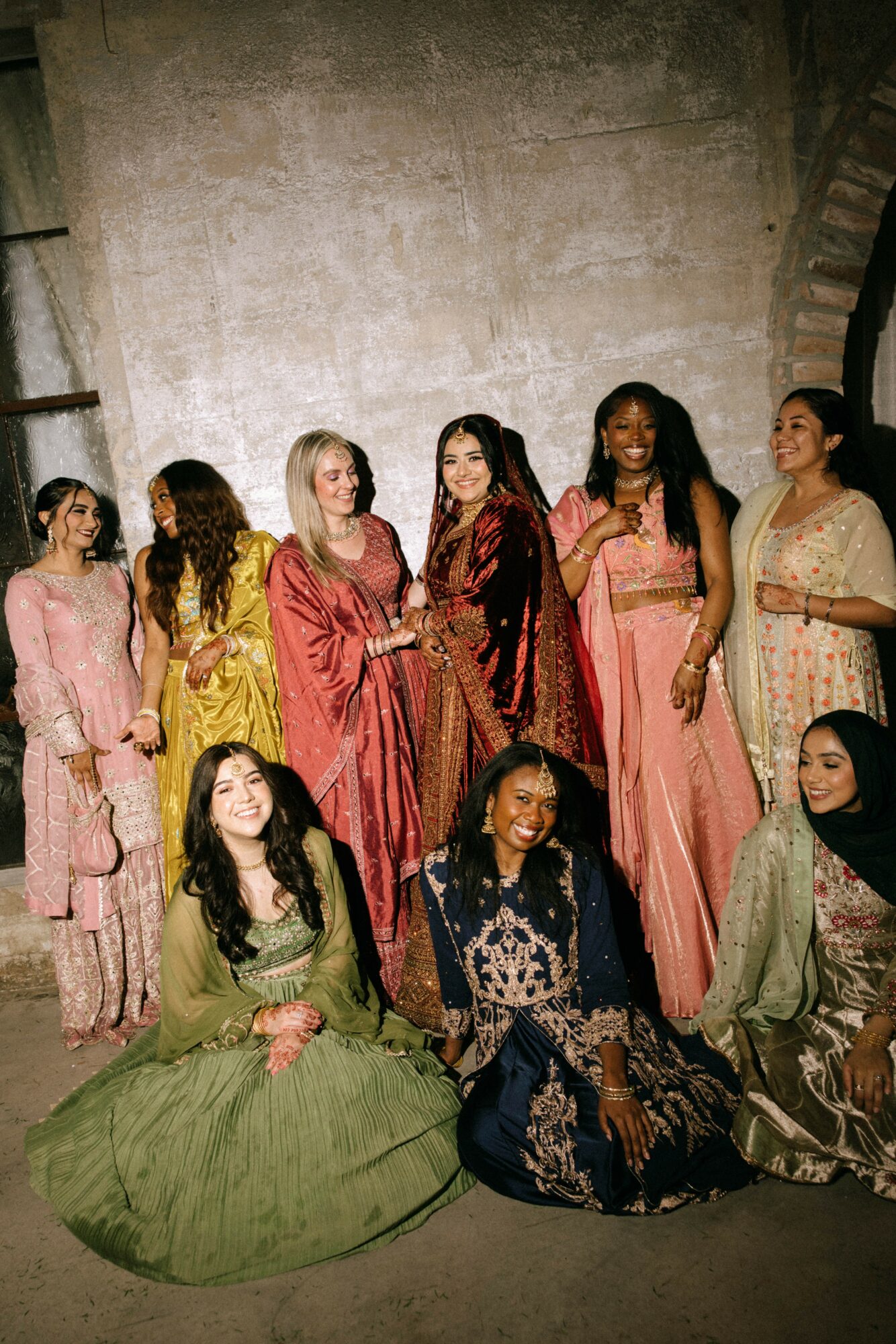 Group of women in colorful dresses standing and sitting against a textured wall, smiling and posing.