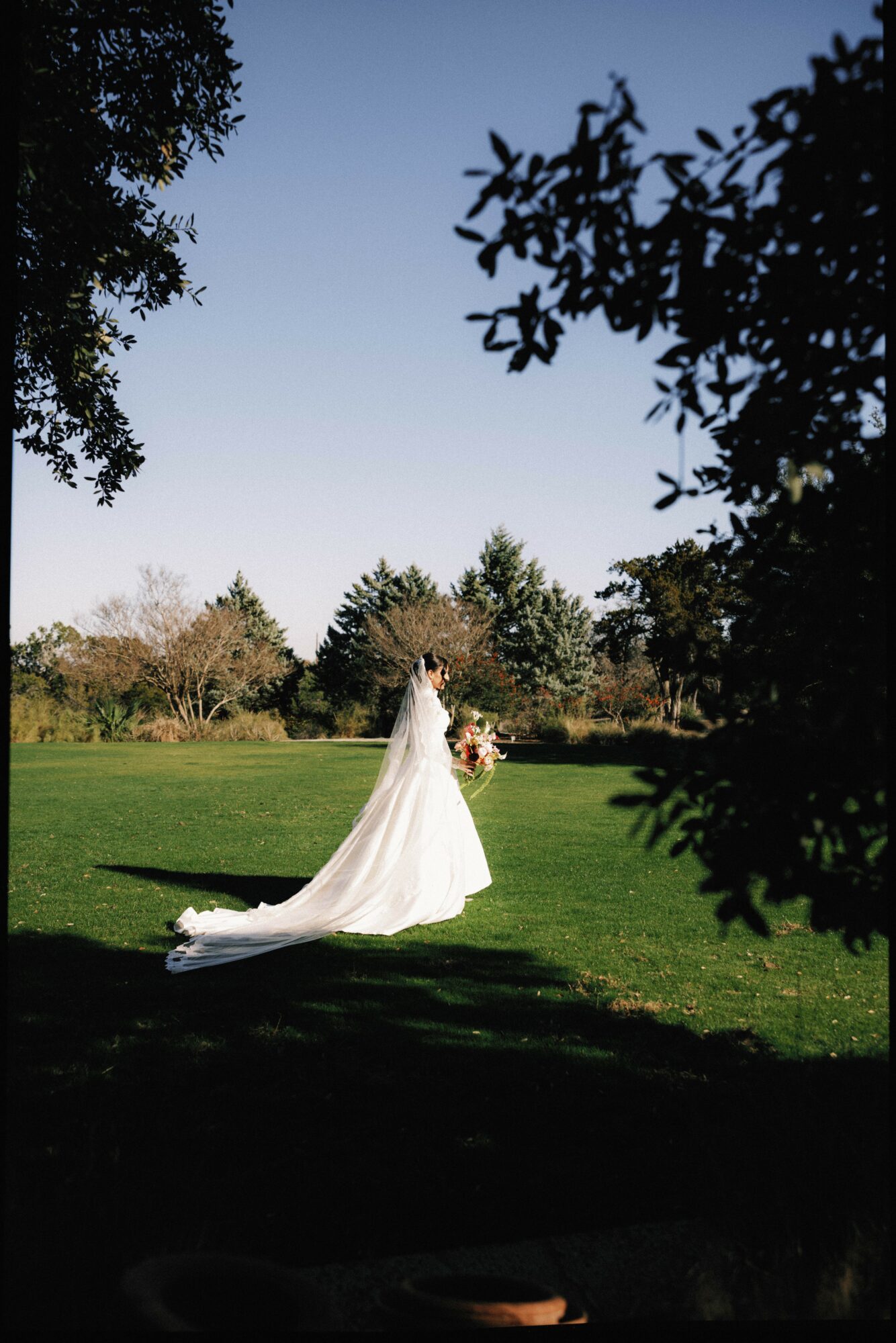 Bride in a white wedding dress holding a bouquet standing on grass with trees and blue sky in background.