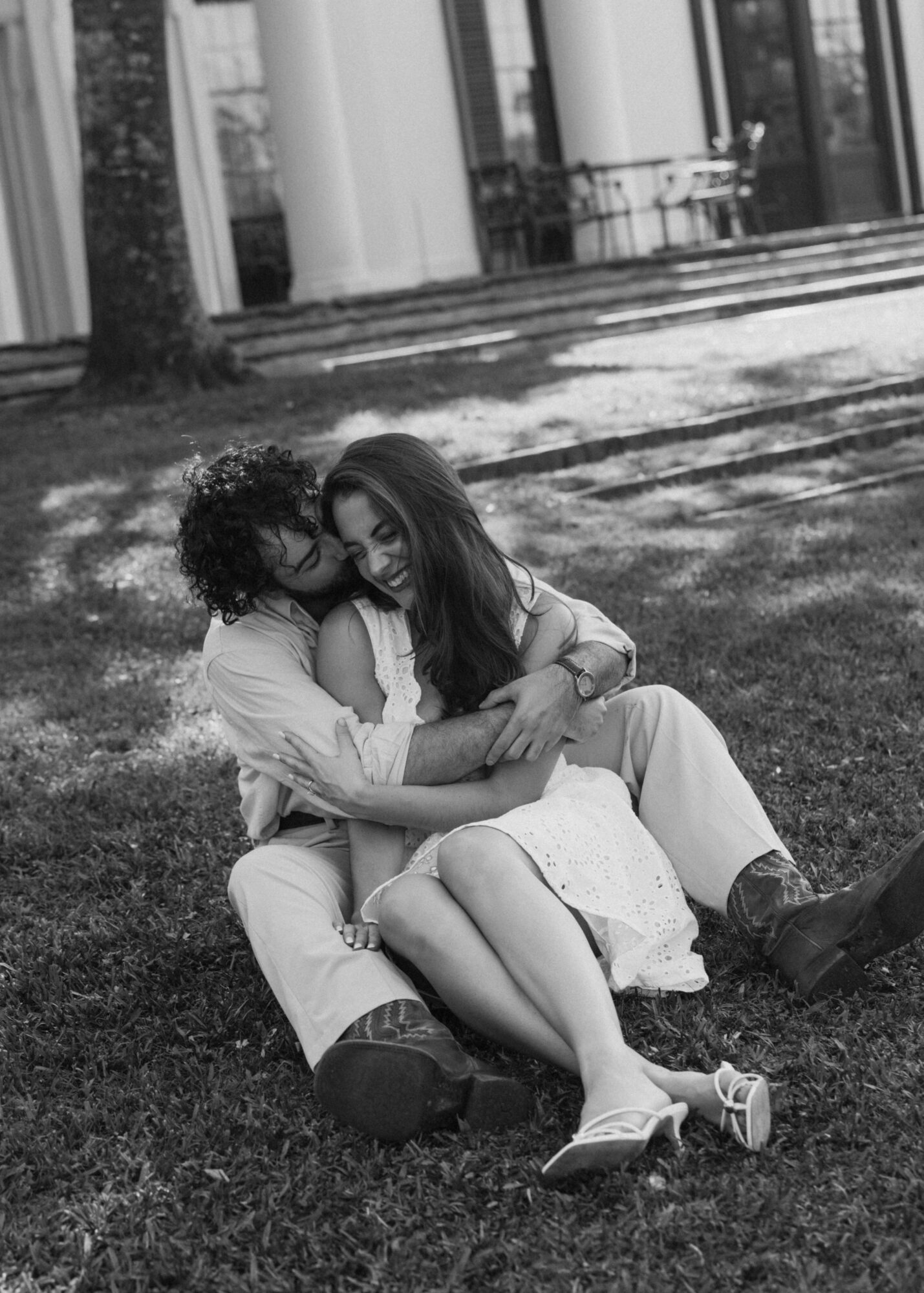 Two people sit on grass, embracing and leaning their heads together outdoors with columns and trees in background.