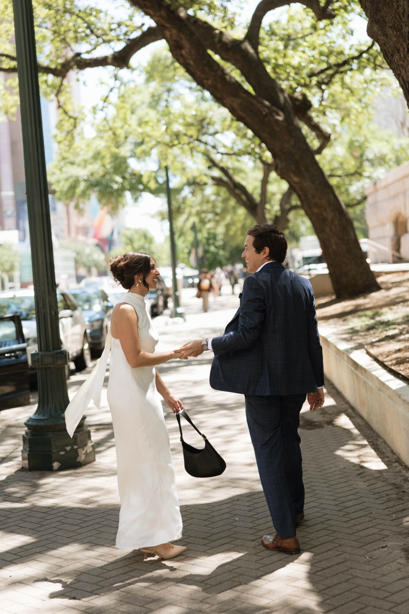 Two people, a woman in a white dress and a man in a dark suit, hold hands on a sunny sidewalk with trees.