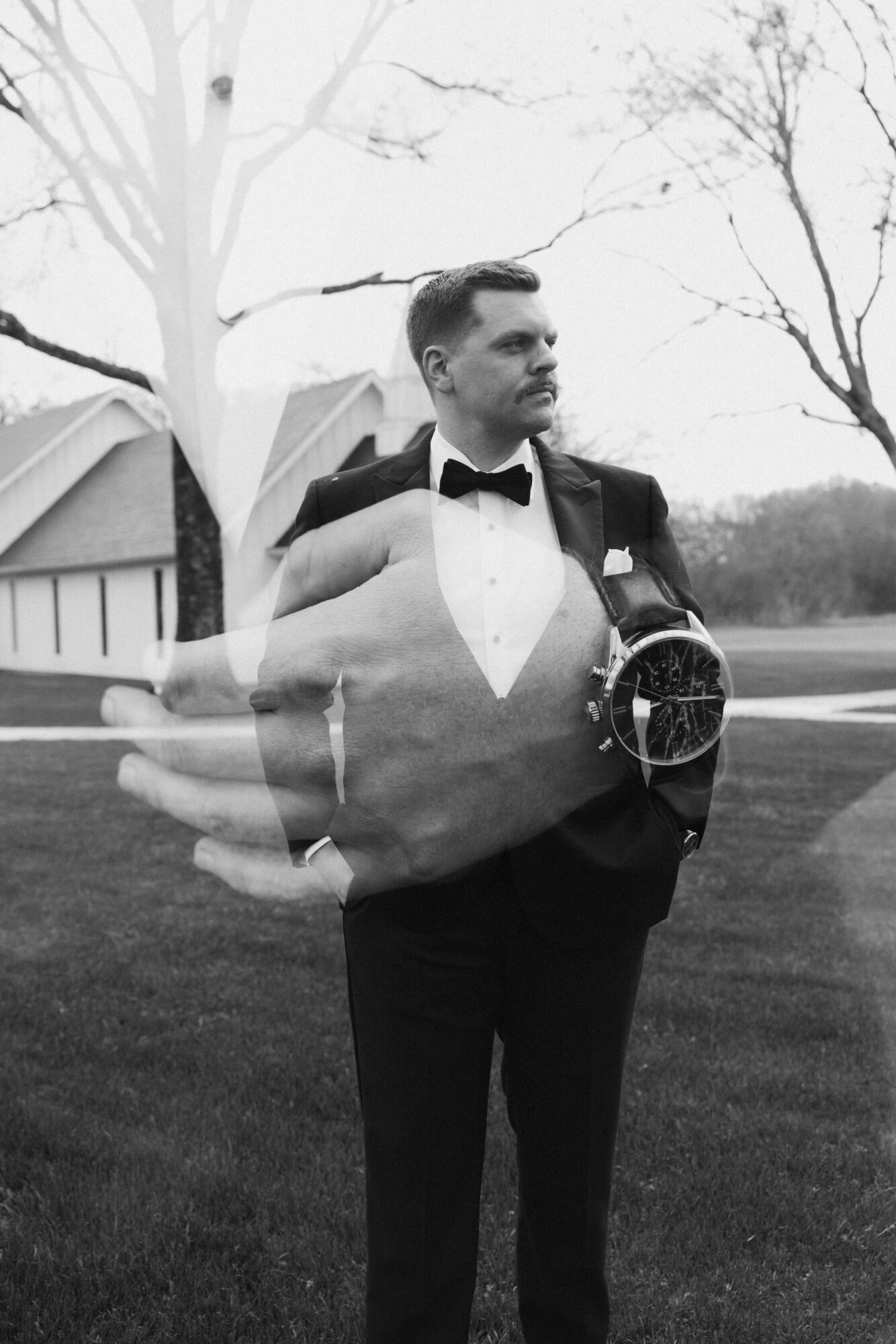Man in tuxedo with bow tie standing outdoors, holding a camera, with trees and a building in background.