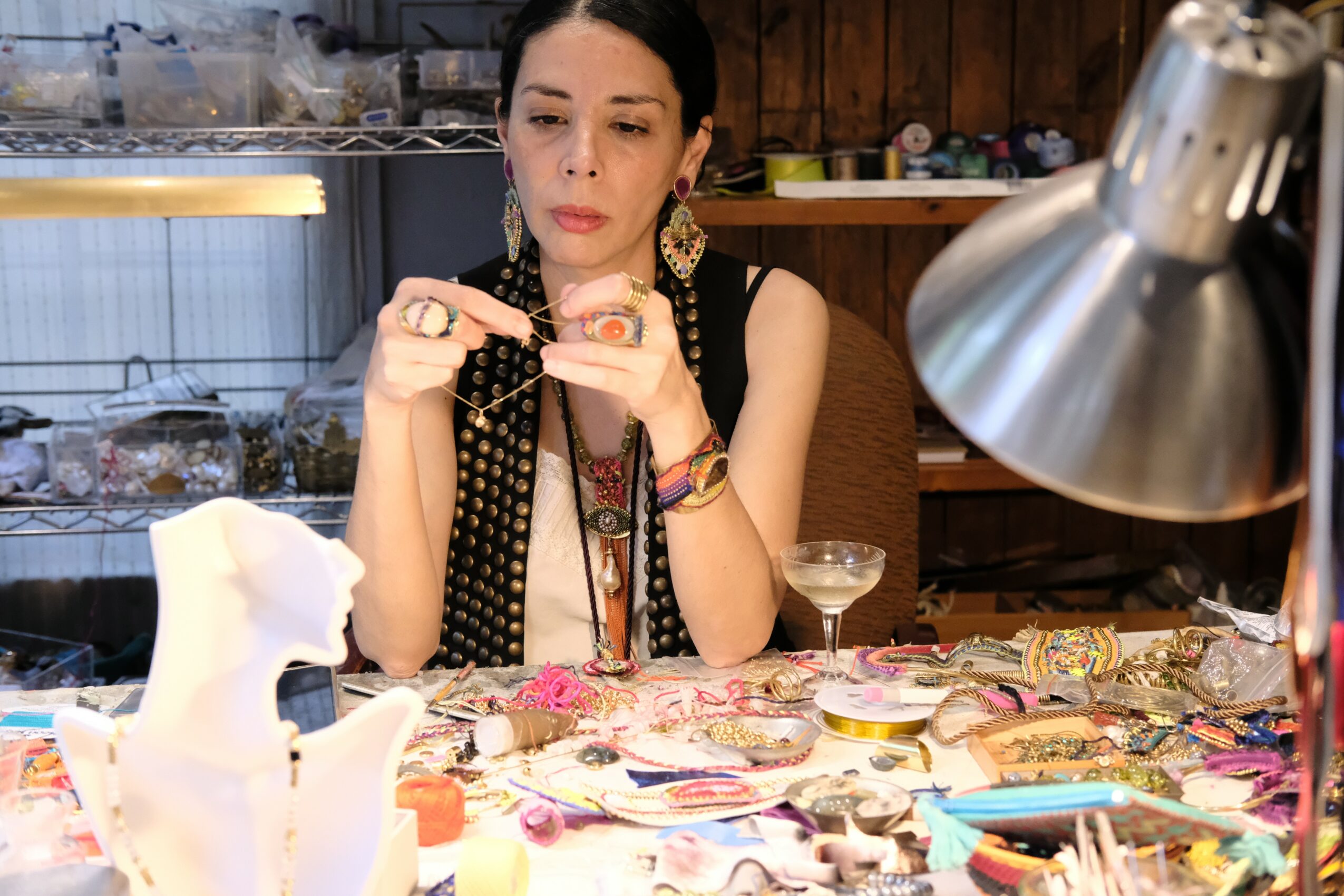 Woman with dark hair and earrings sits at a cluttered table, holding a small object, with a lamp overhead and a glass nearby.