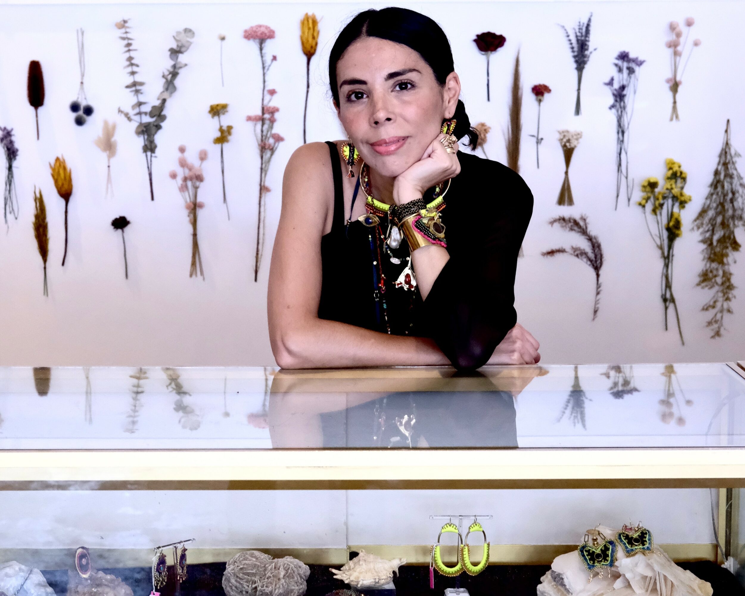 Woman with dark hair and colorful jewelry leaning on a glass display case, floral wall behind her, smiling.