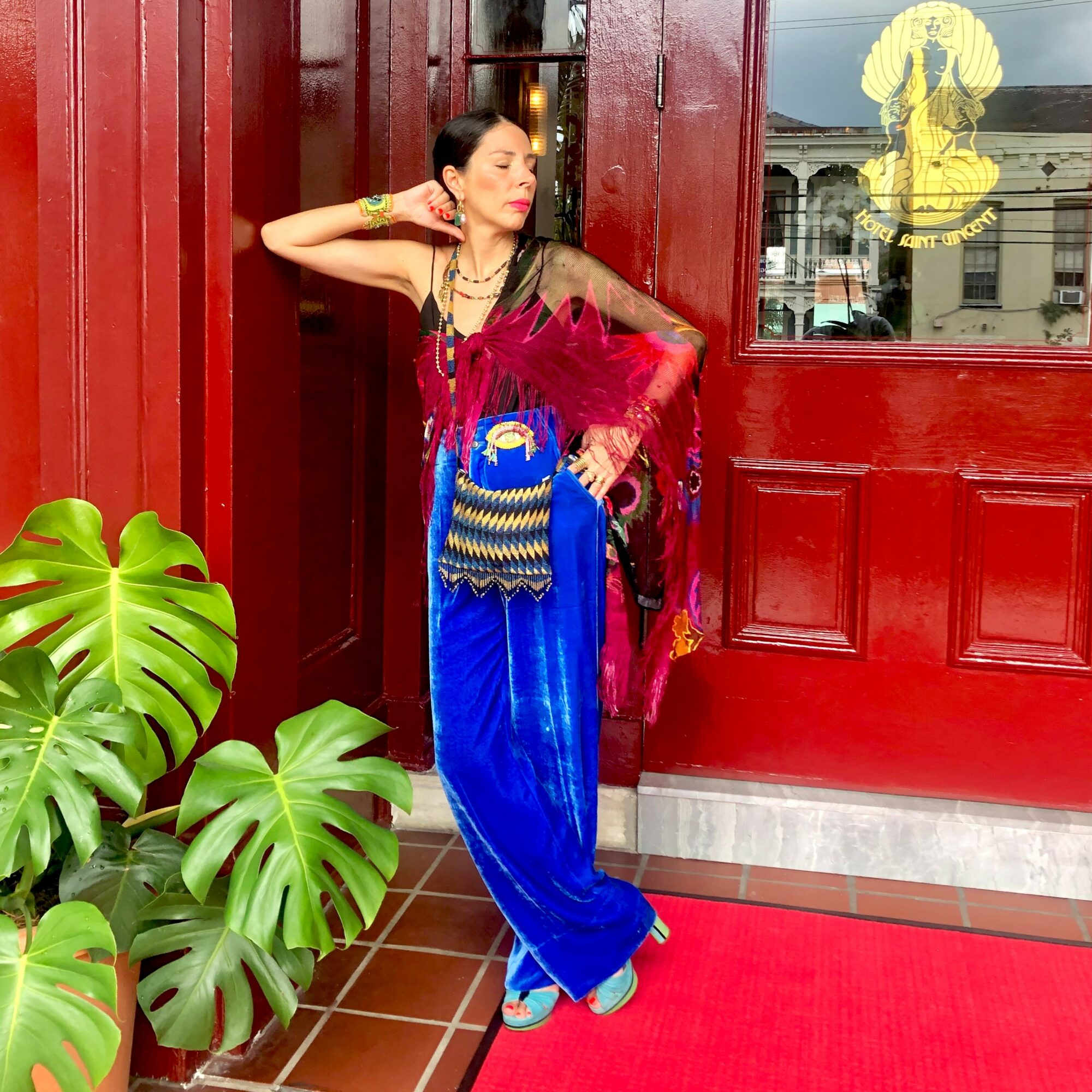 Woman in colorful traditional attire standing by a red door with a window, plants nearby, looking to the side.