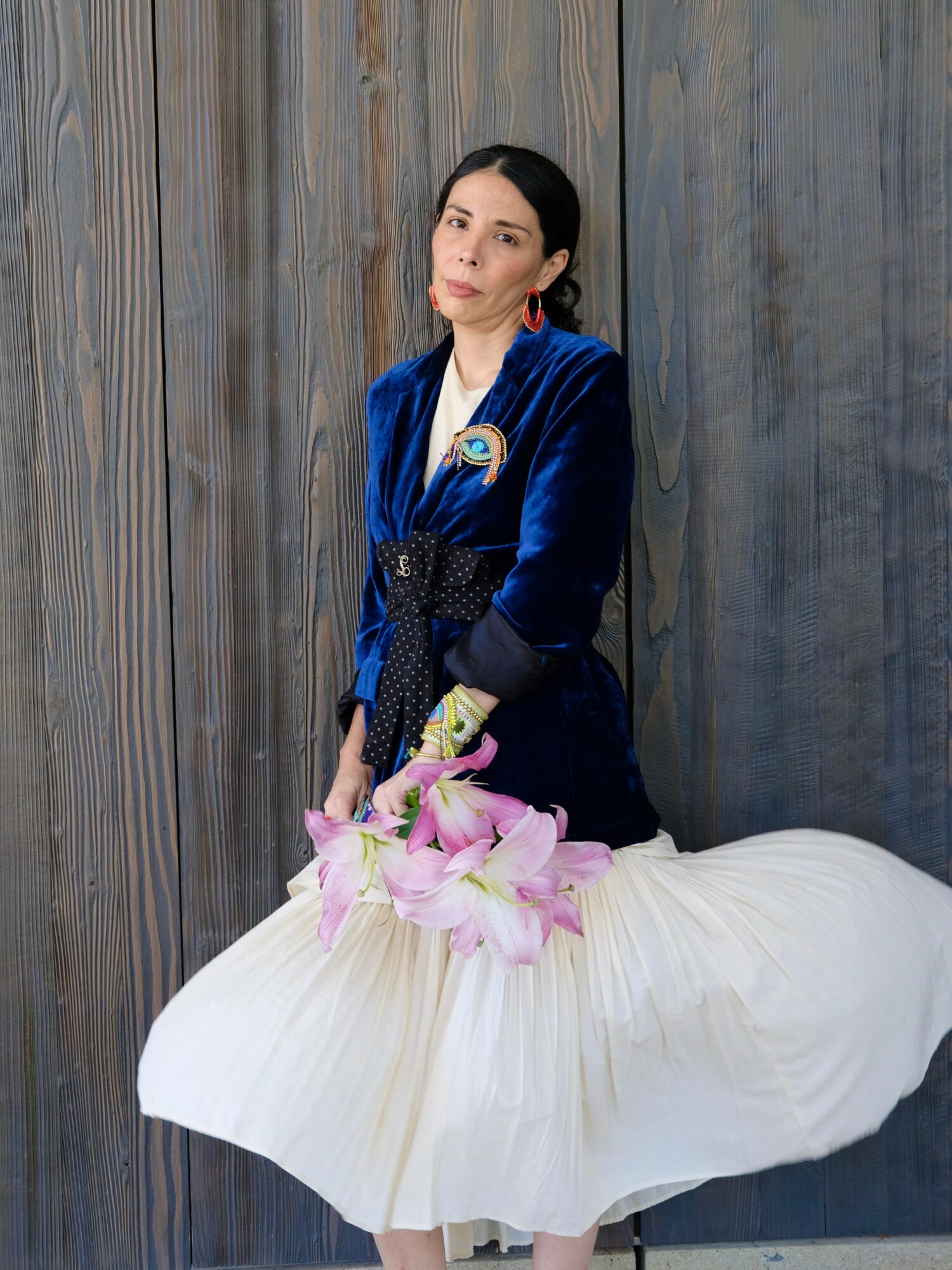 Woman in blue velvet dress holding pink flowers, standing against wooden wall, with a white pleated skirt in front.